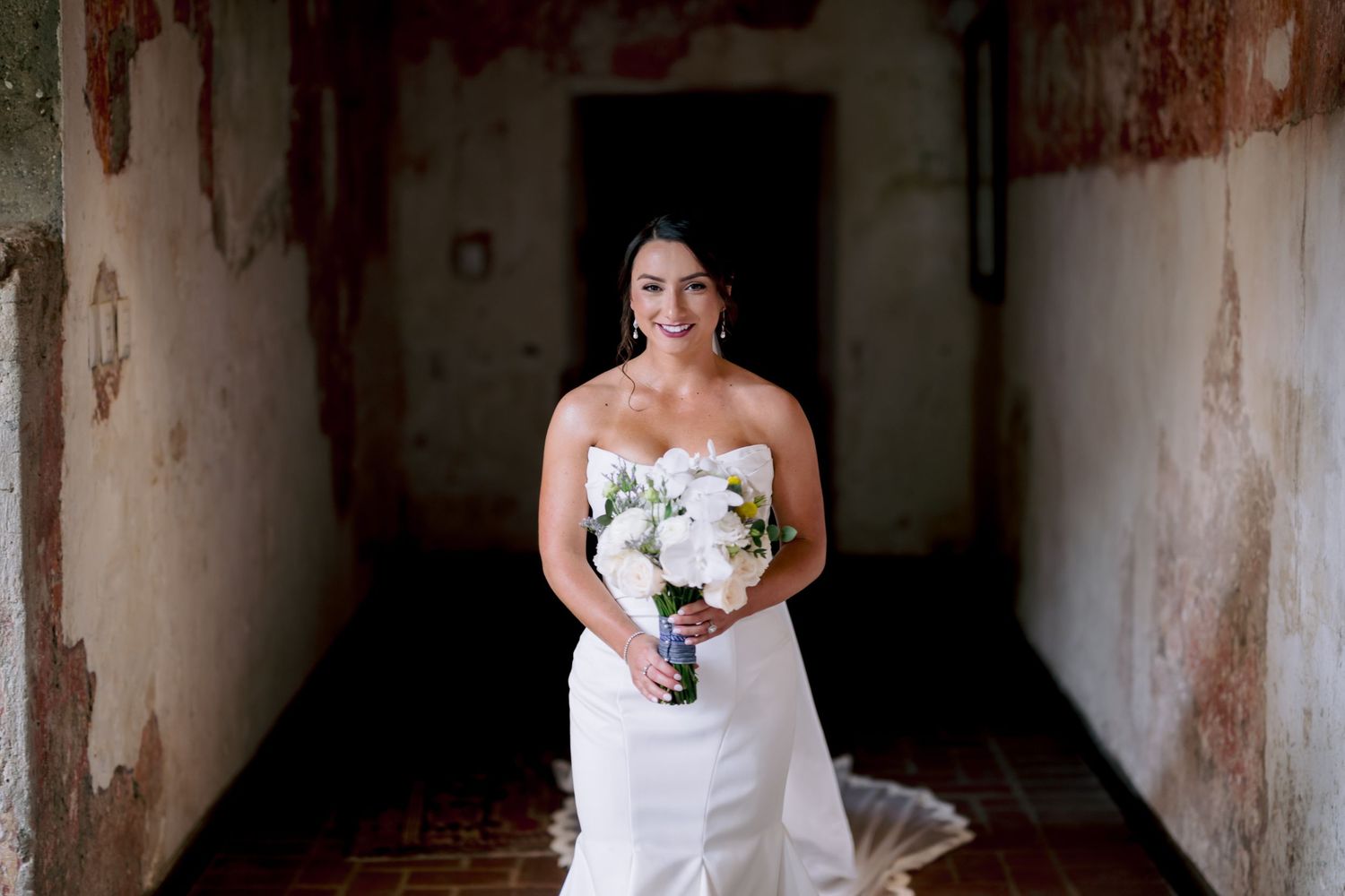 A bride in a fitted white dress holds a bouquet while standing in a historic corridor.