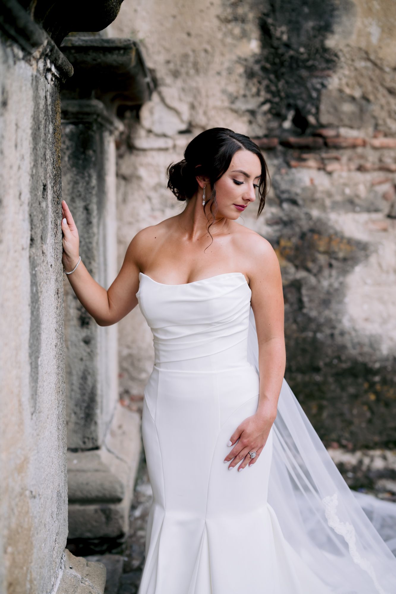 A bride in a fitted strapless gown poses elegantly against a weathered stone wall.