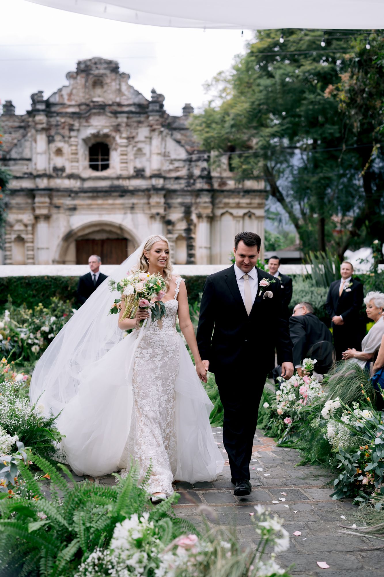 Newlyweds walking down garden path with historic church facade and string lights overhead.