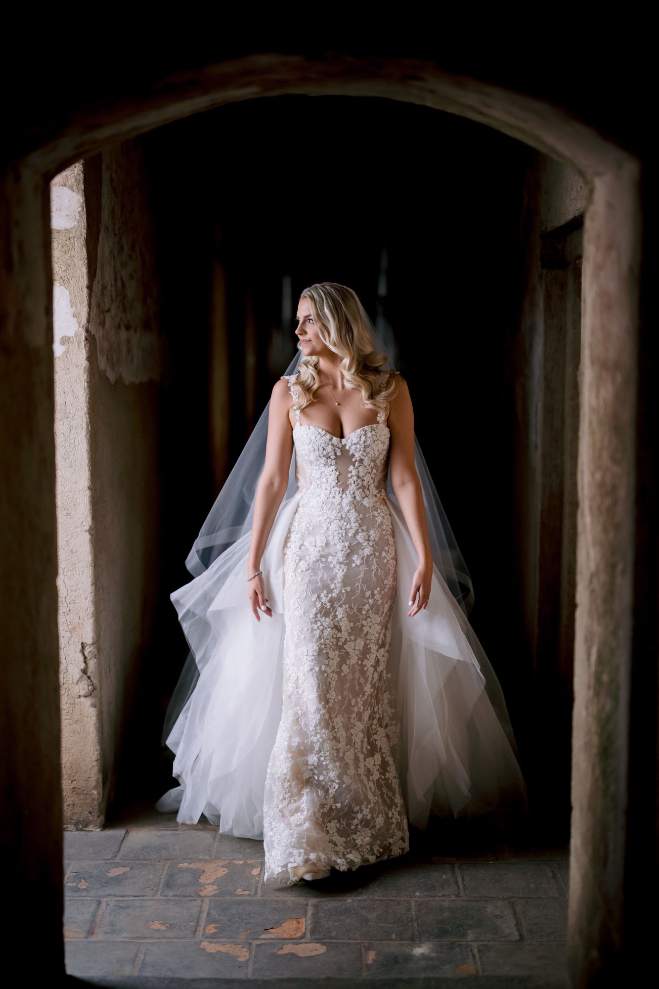 Elegant bride in fitted lace gown stands gracefully in stone archway with flowing veil.