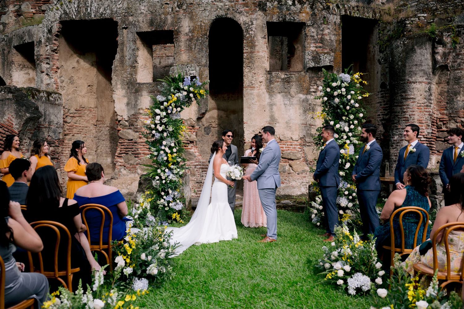 Outdoor wedding ceremony in ancient ruins decorated with floral arches and wooden chairs.