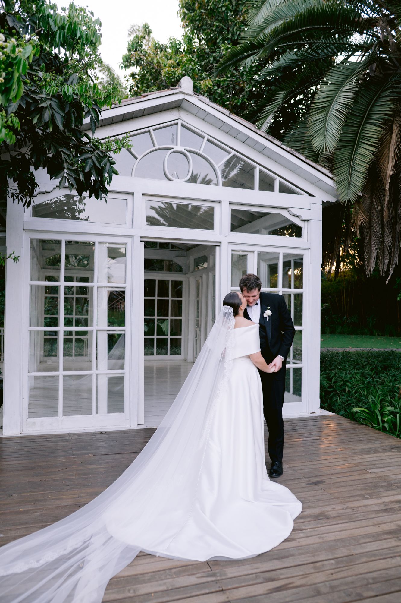Bride in classic white gown and groom pose beside elegant glass conservatory in garden setting.