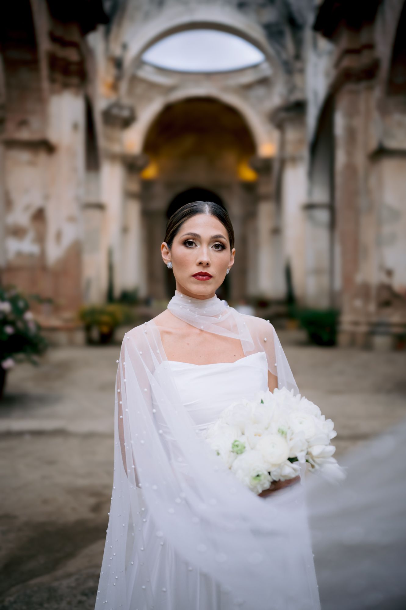 Dramatic bridal portrait showcasing pearl-adorned cape and classic updo against church architecture.