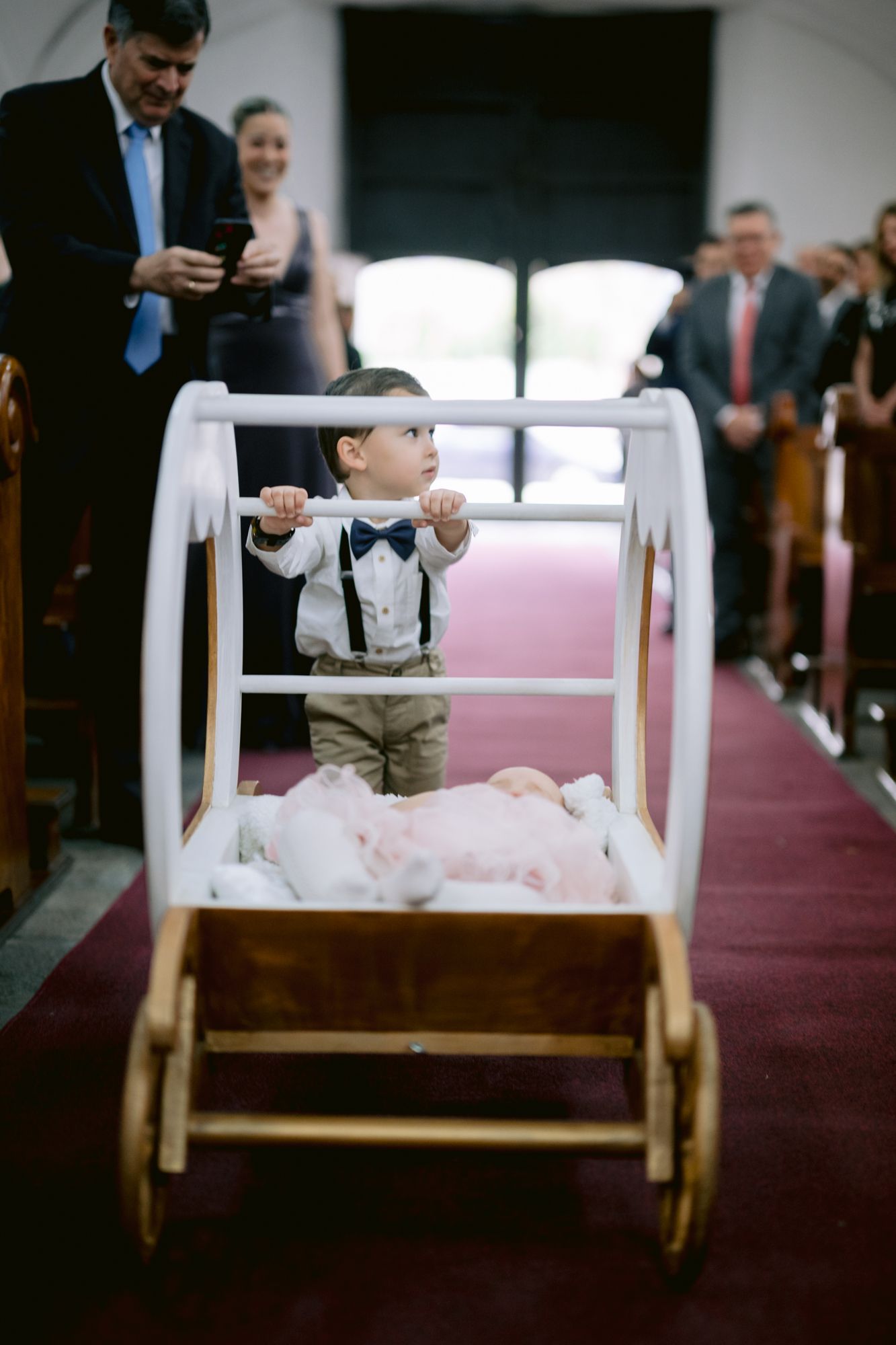 Small child in suspenders pushing decorative white wagon down church aisle.