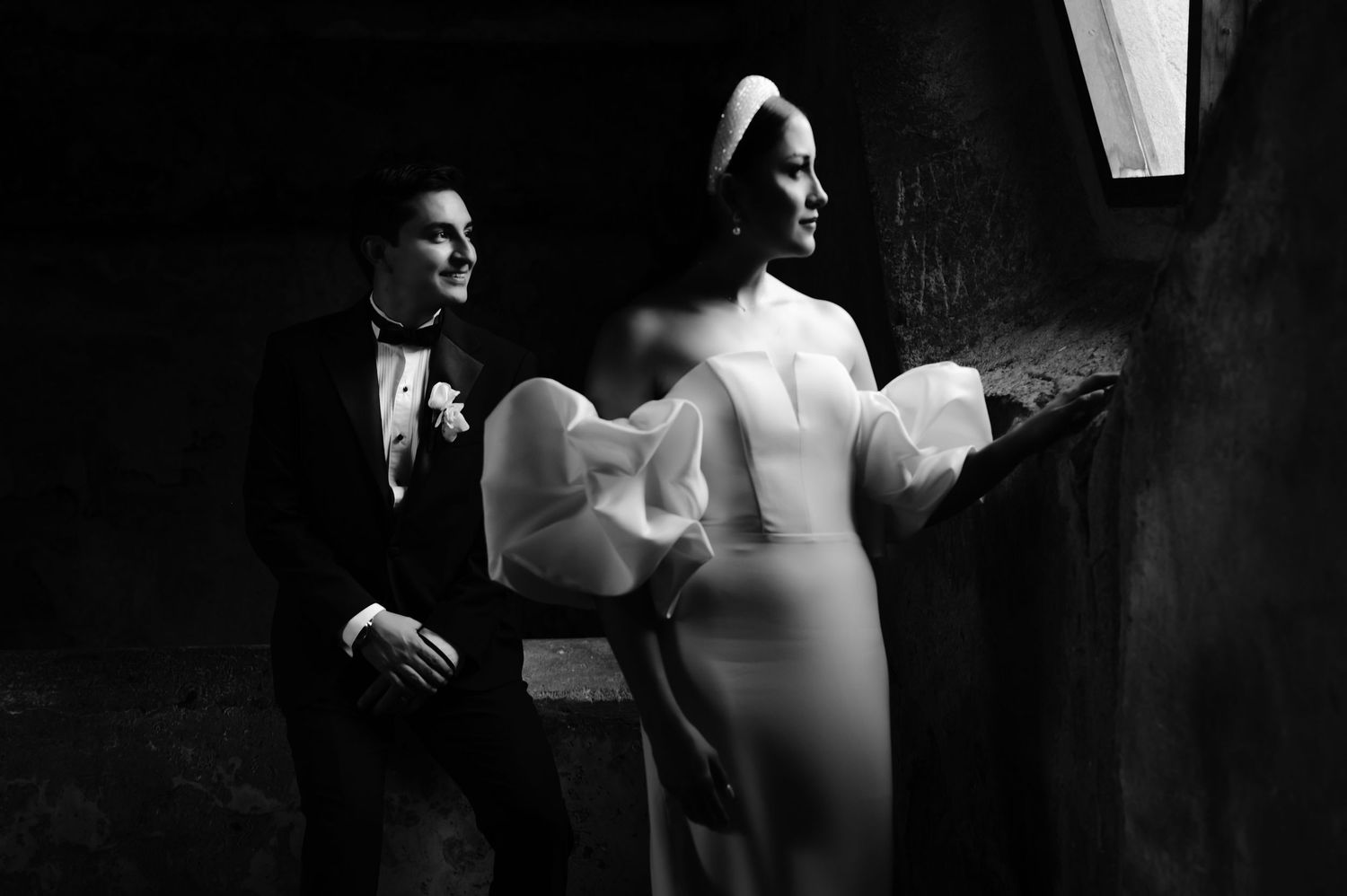 Artistic black and white portrait of wedding couple in moody lighting against stone wall.
