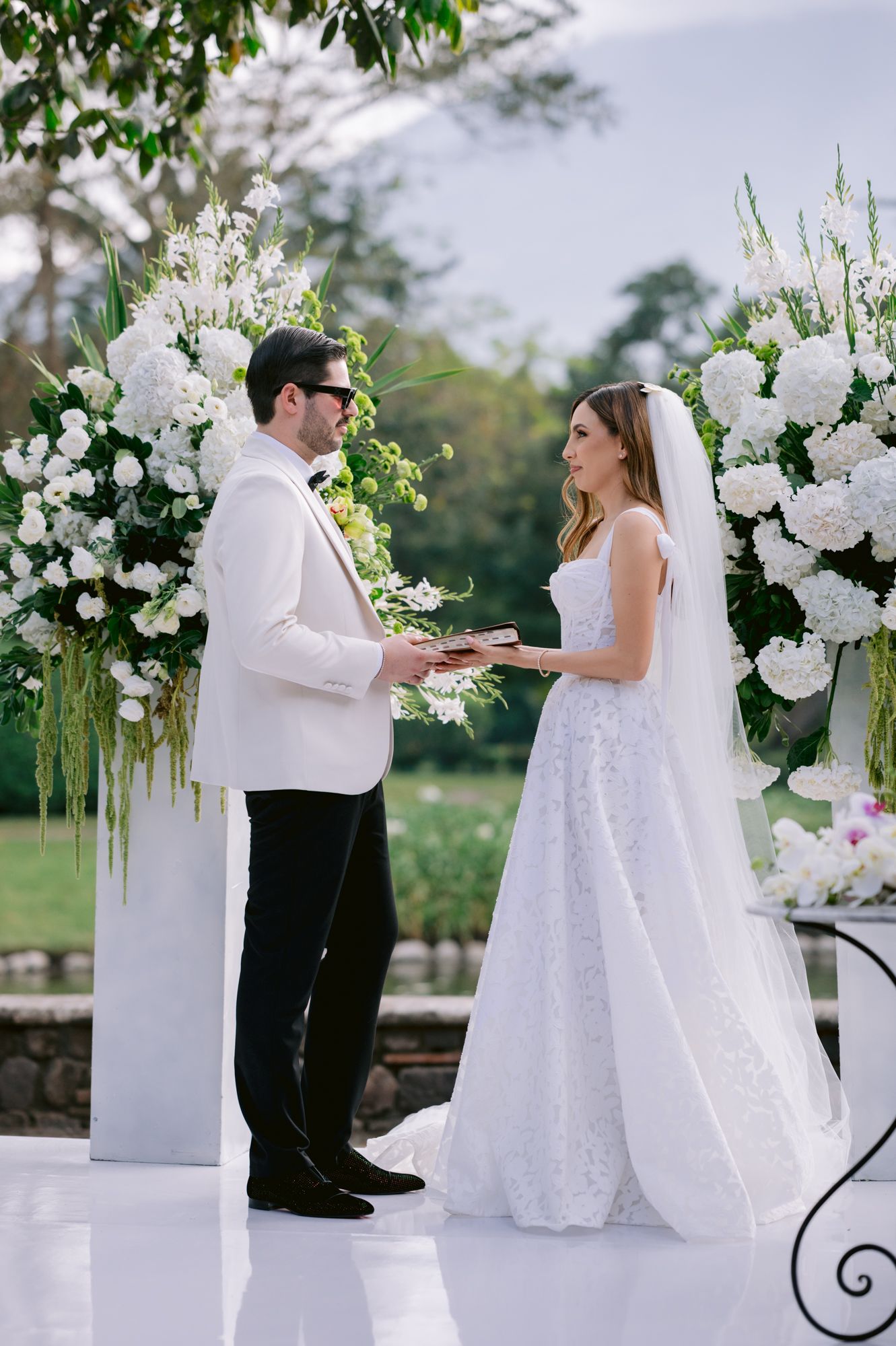 Couple exchanging vows during outdoor wedding ceremony surrounded by white floral arrangements and columns.