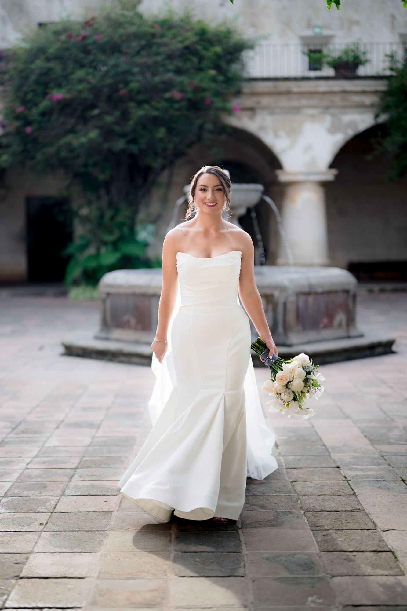 A stunning bride poses in a fitted white wedding gown against a Spanish colonial courtyard setting.