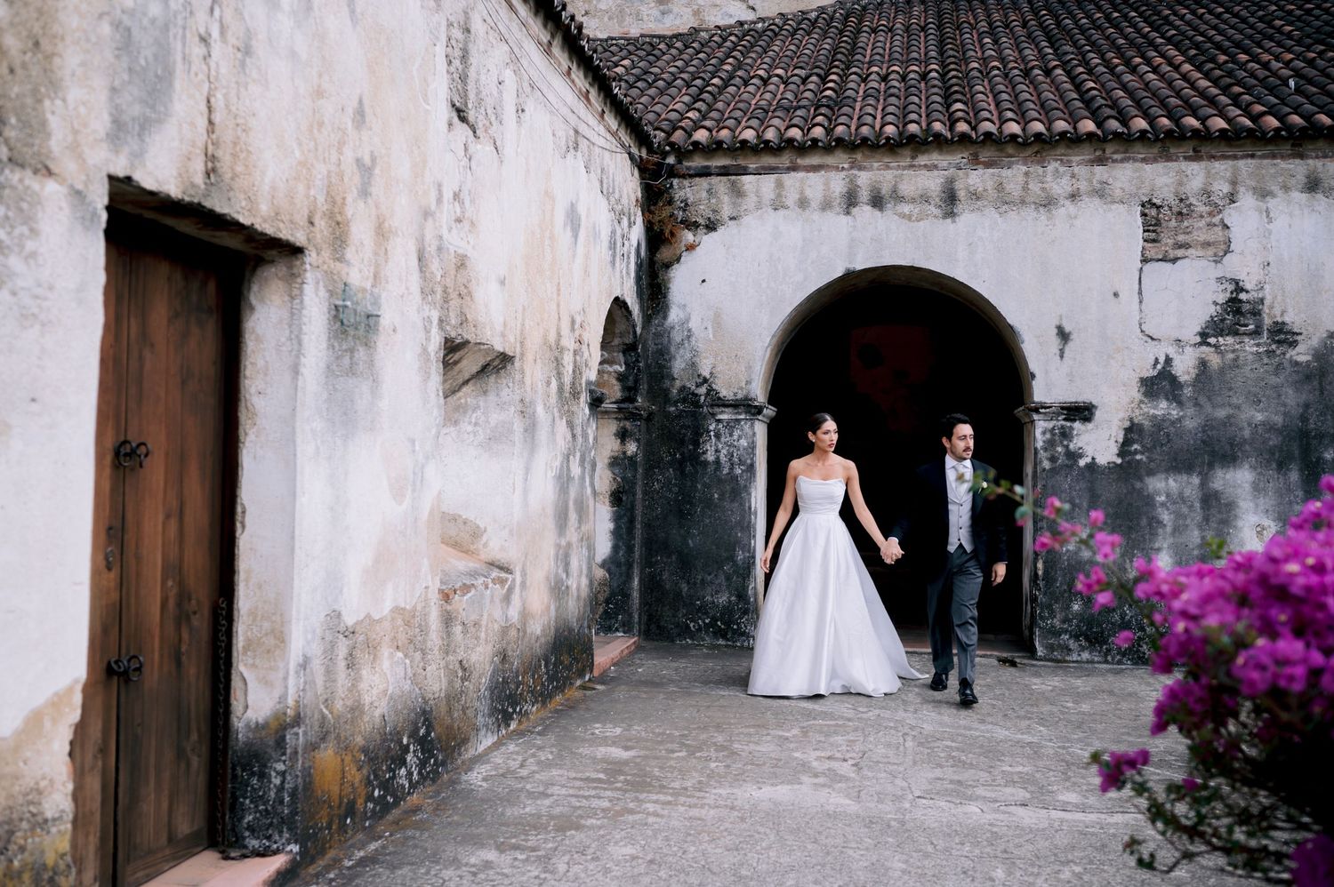 A couple walks through an old stone archway adorned with vibrant pink bougainvillea flowers.