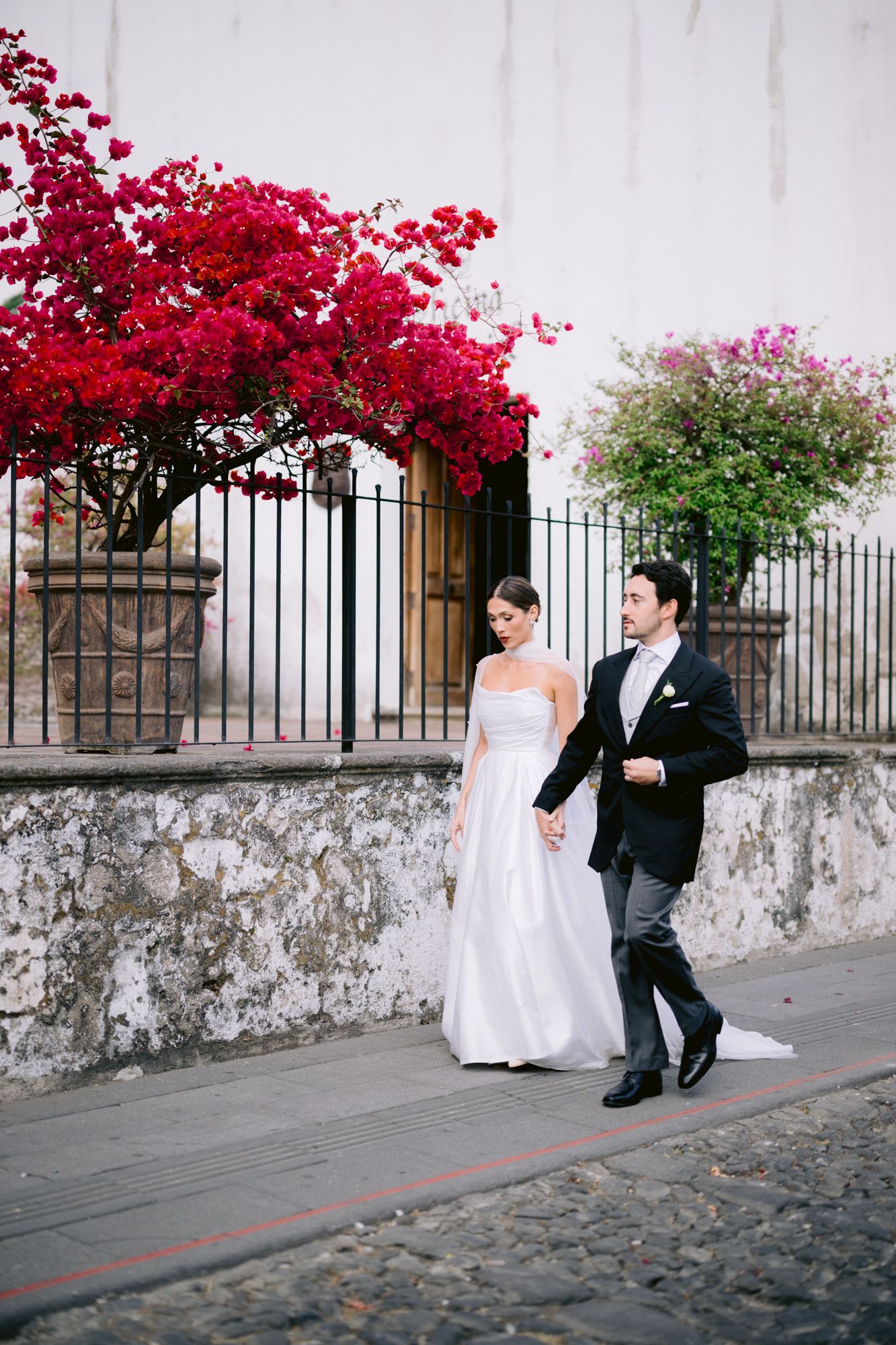 A wedding couple walks along a stone wall adorned with vibrant pink bougainvillea flowers in an historic setting.
