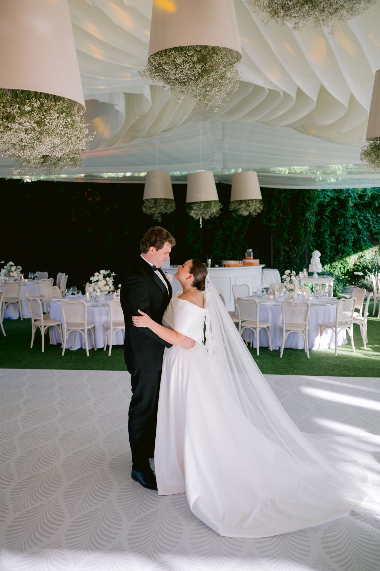 An elegant wedding couple dances on a patterned floor beneath decorative white ceiling drapes.