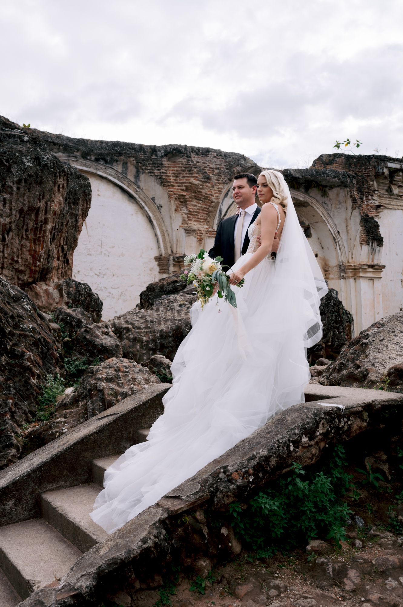 A wedding couple poses on ancient stone steps against white arched ruins in a dramatic architectural setting.