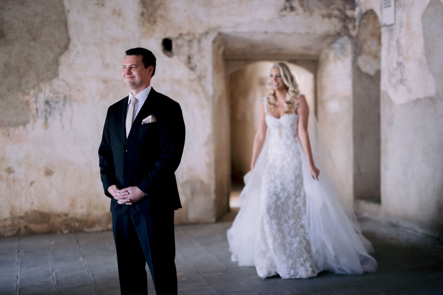 Wedding first look with groom waiting as bride stands in background of stone corridor.