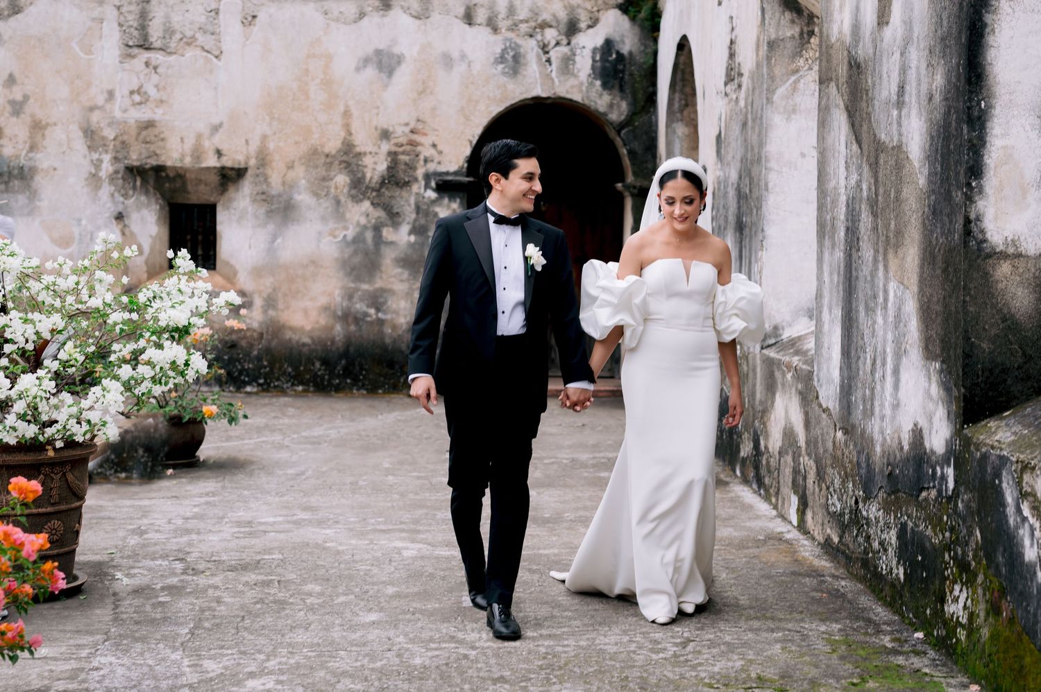 A newlywed couple walks together through a rustic courtyard with weathered walls and potted flowers.