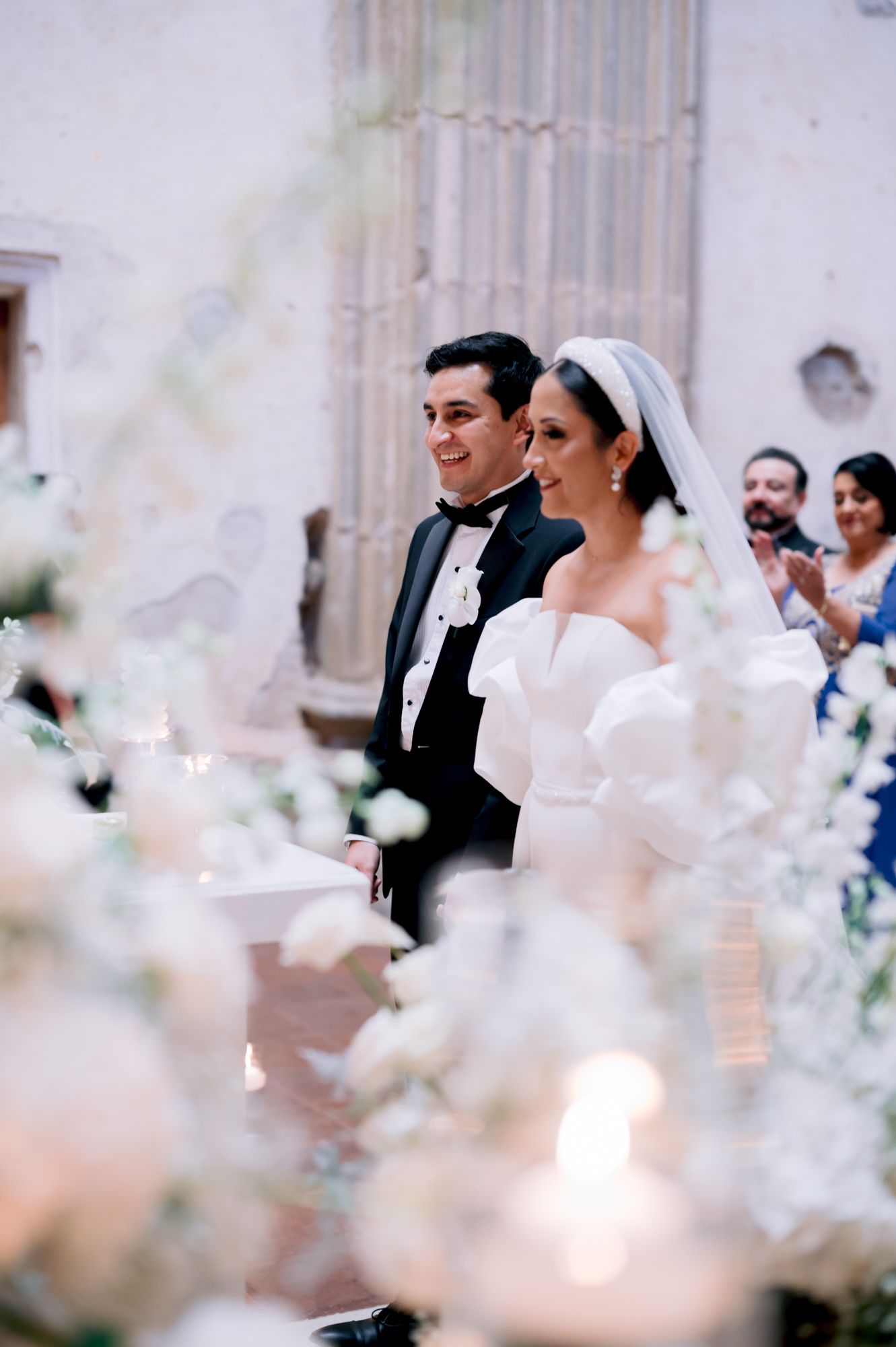 Wedding ceremony with soft focus floral decorations in foreground against stone walls.