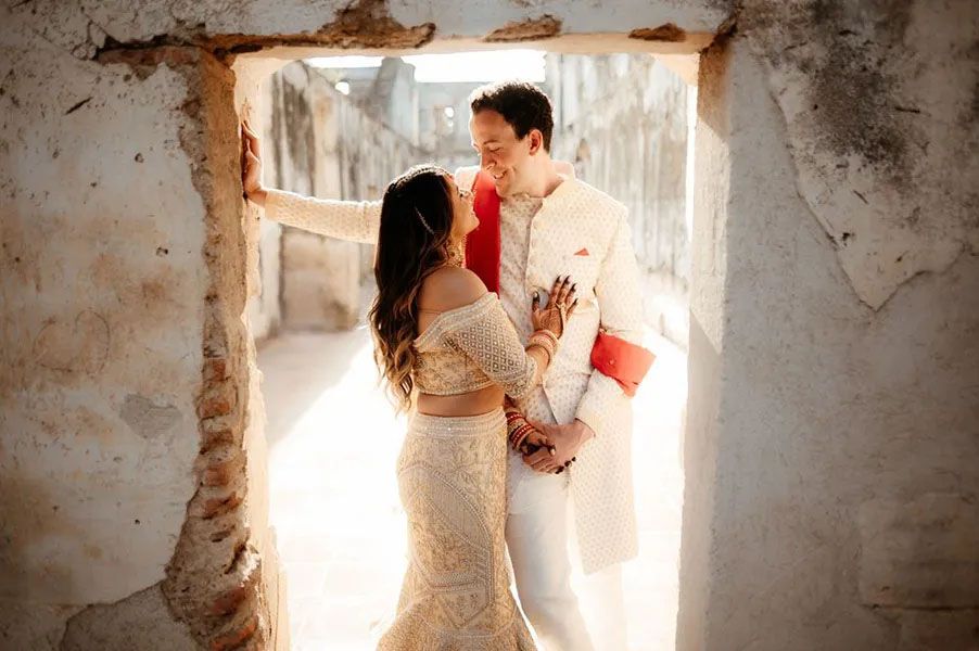 A romantic wedding couple embraces in a sunlit stone doorway wearing elegant white attire.