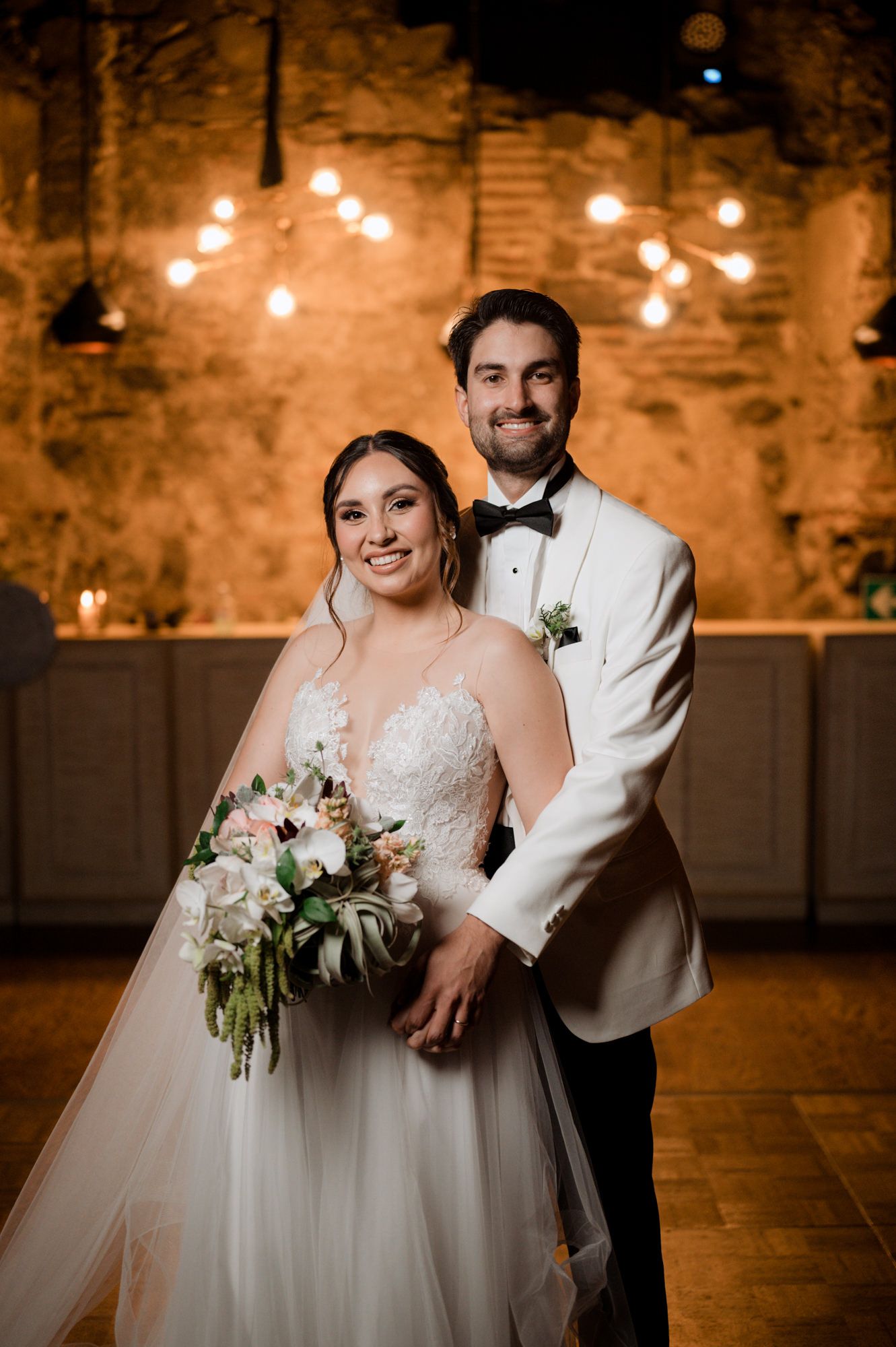Wedding portrait in a warm-lit venue with stone walls and hardwood floors.
