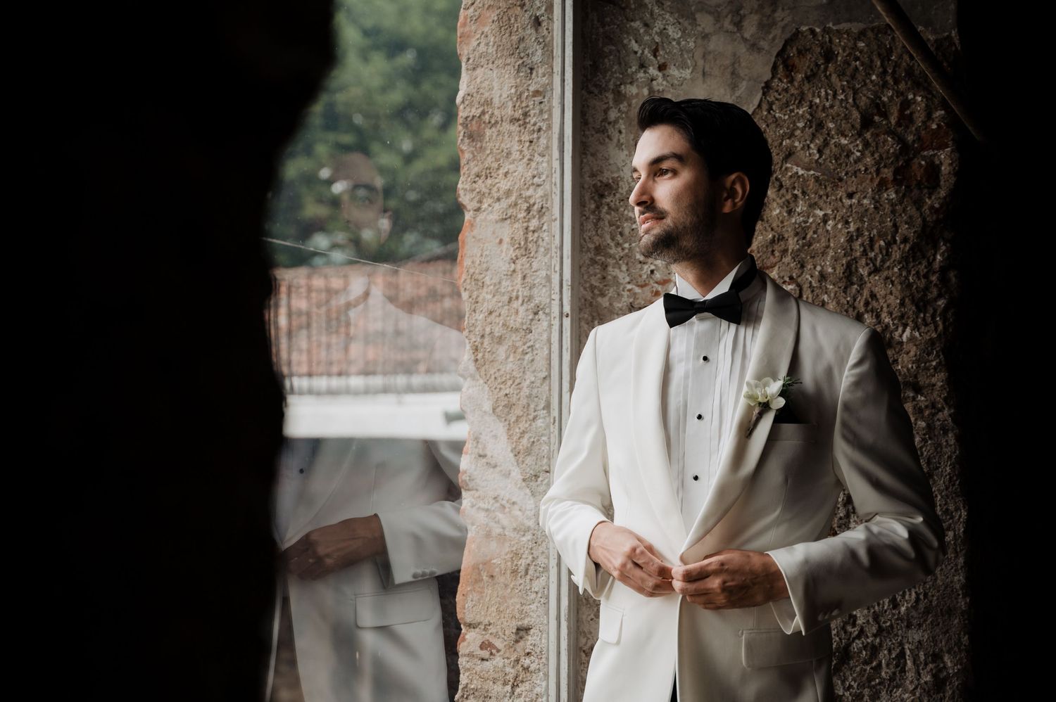 Groom in white tuxedo jacket poses thoughtfully by stone archway in classic portrait.