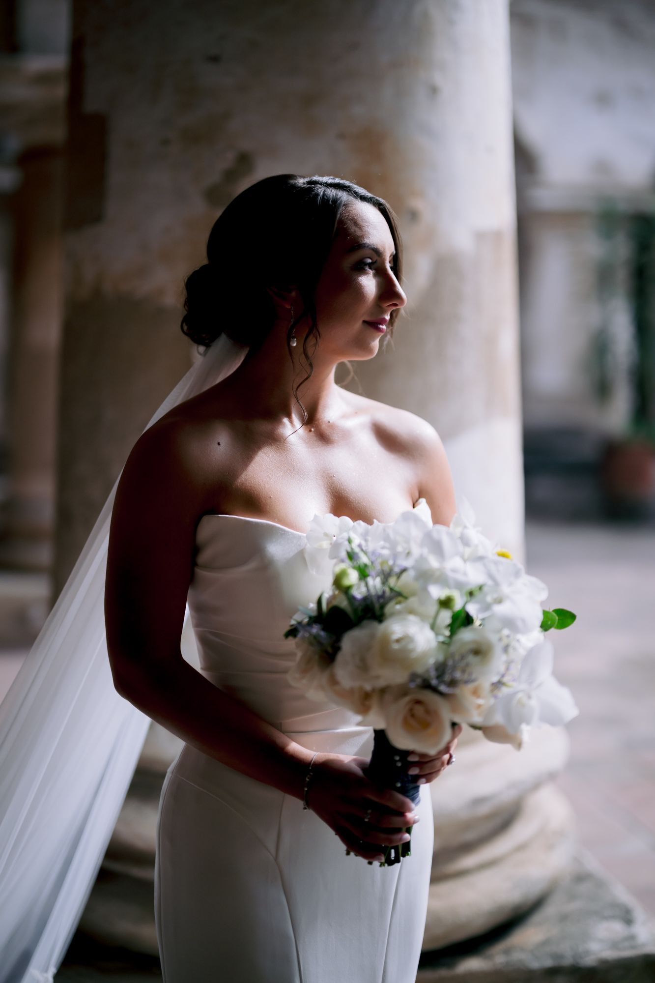 Elegant bridal portrait with white bouquet against a classic stone column in dramatic lighting.