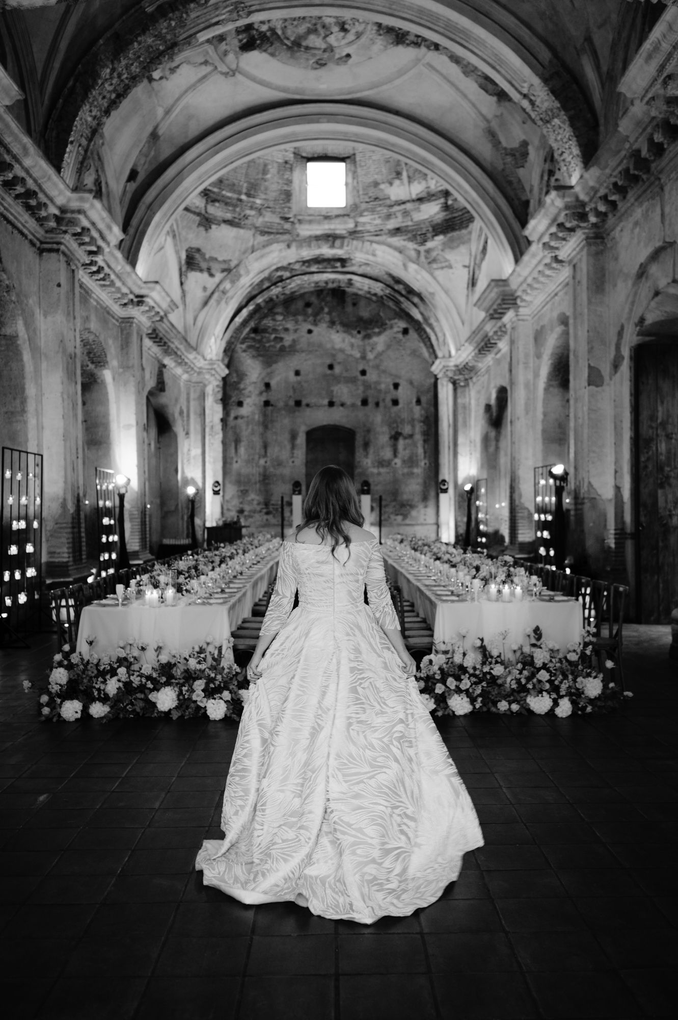 Bride in flowing white gown stands in ornate church hall with vaulted ceilings and reception tables.