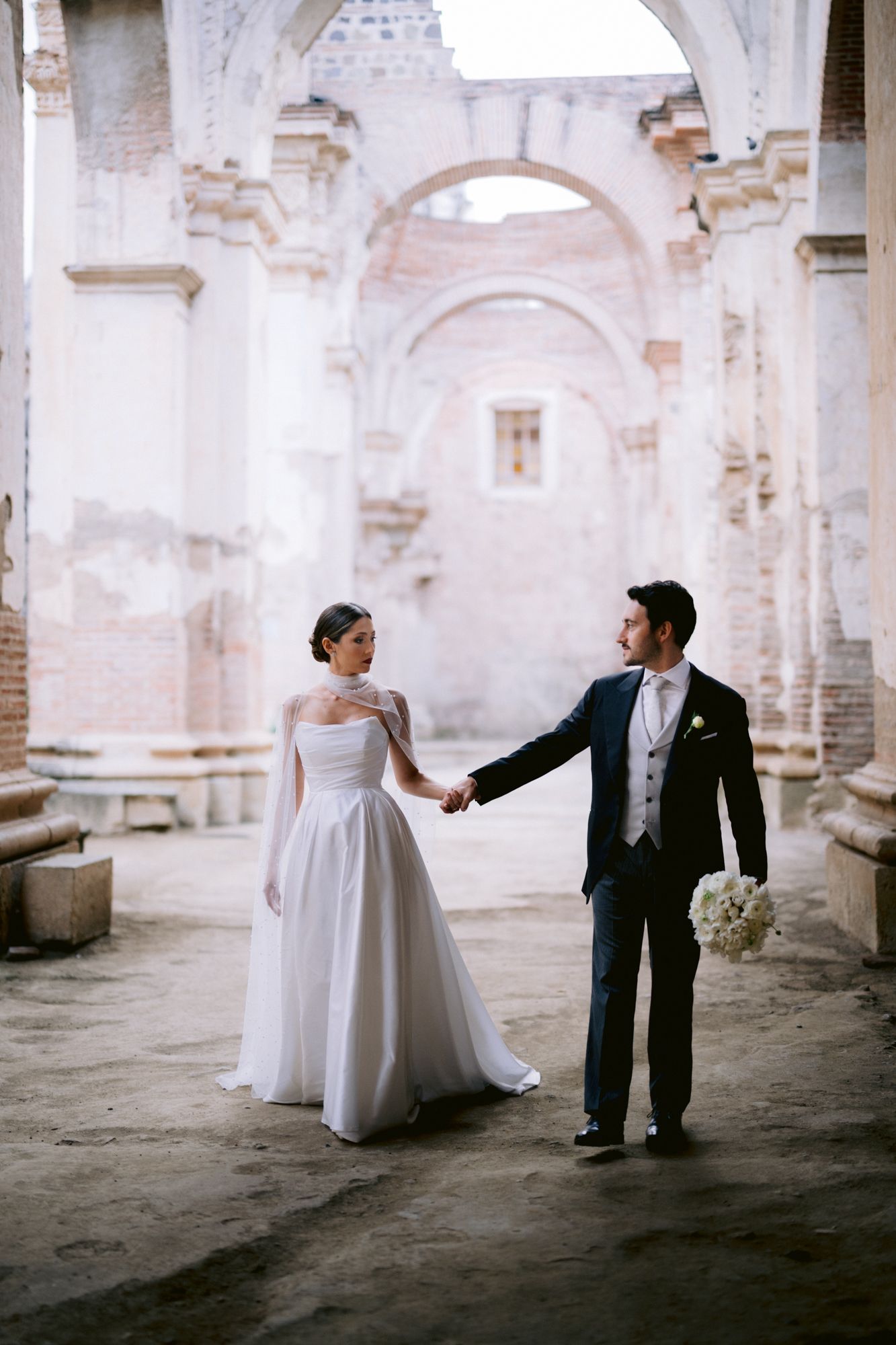 Bride and groom hold hands while walking through ornate stone hallway with arched ceilings.