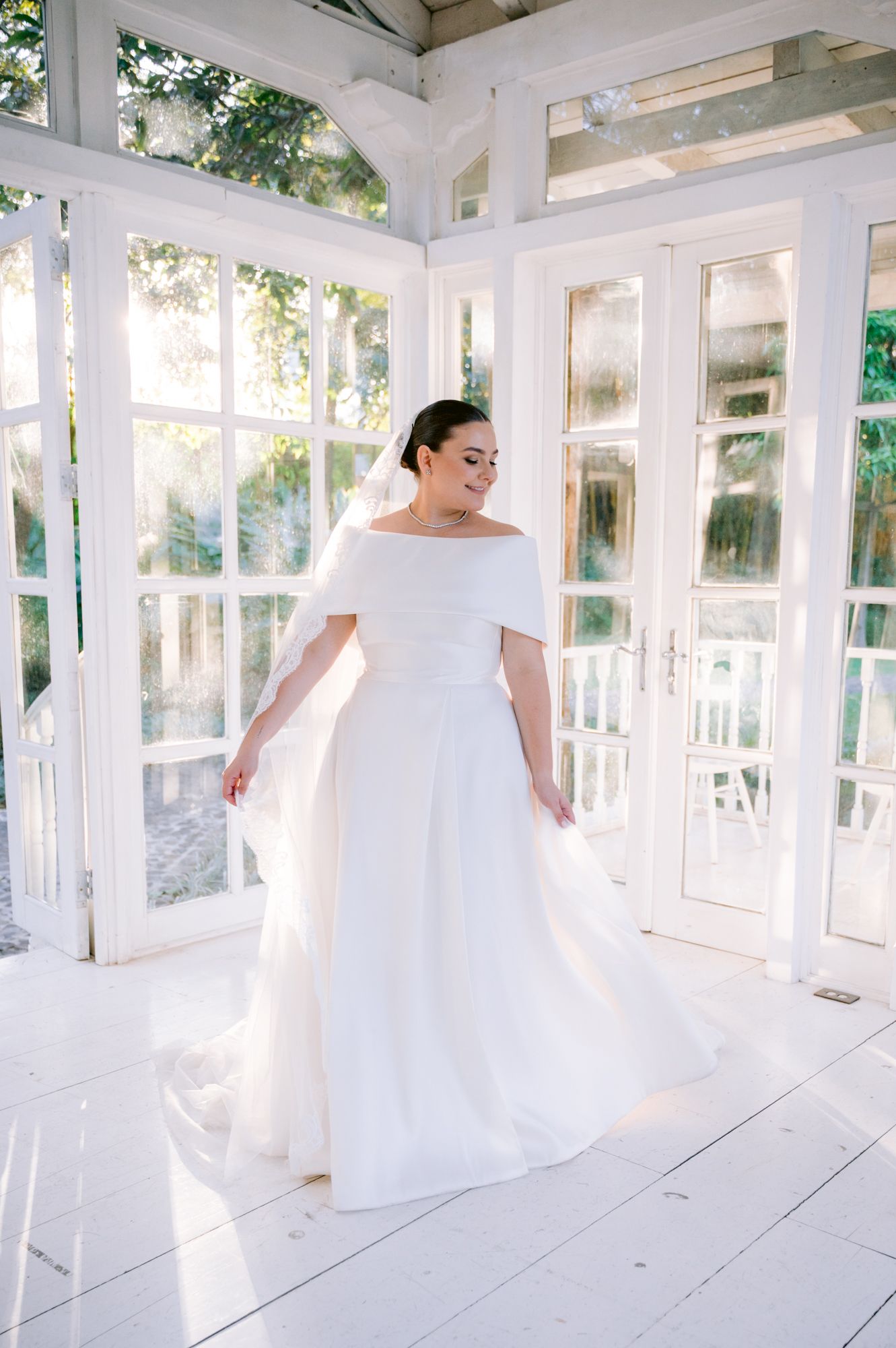 Bride in an elegant off-shoulder wedding gown twirls in a bright white conservatory setting.