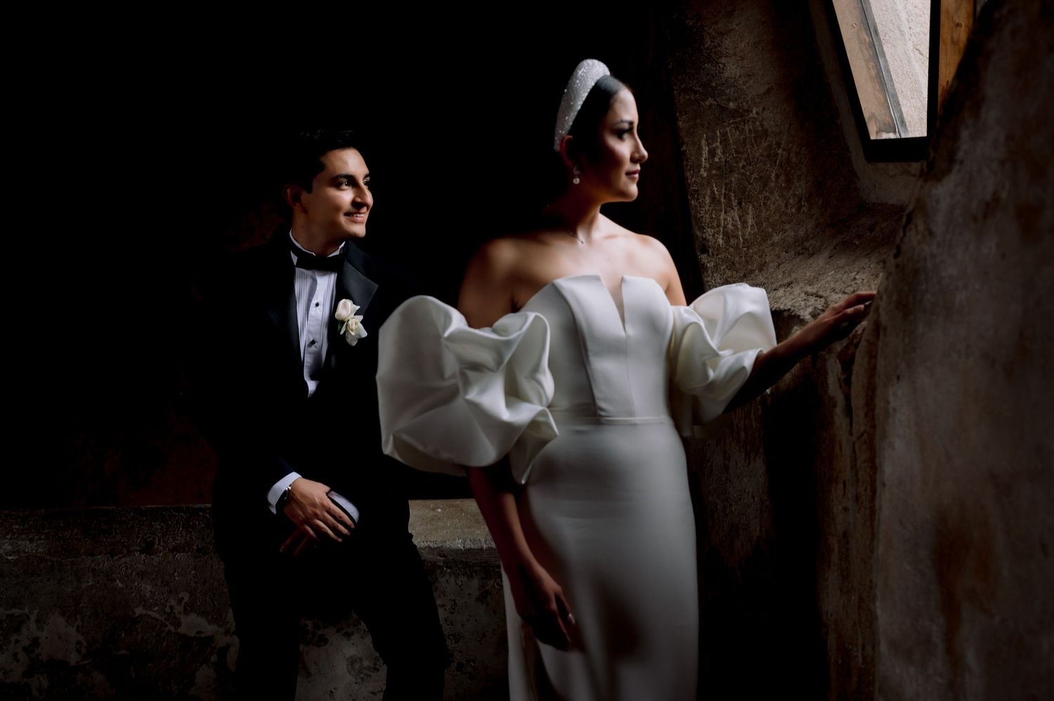 Wedding couple in formal attire pose dramatically in moody lighting against stone wall background.