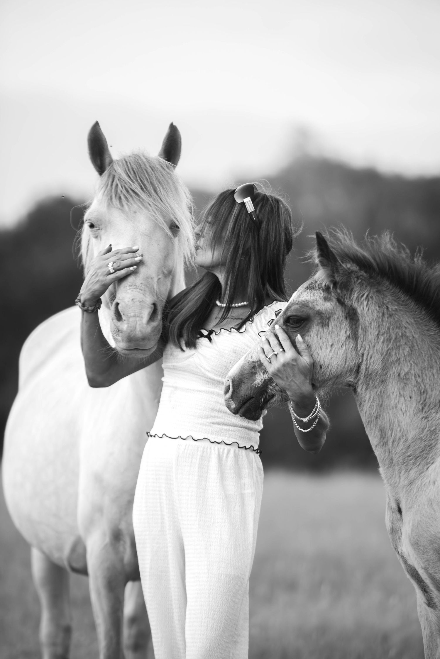 A woman in white dress gently holds the faces of a white horse and a foal in a field.
