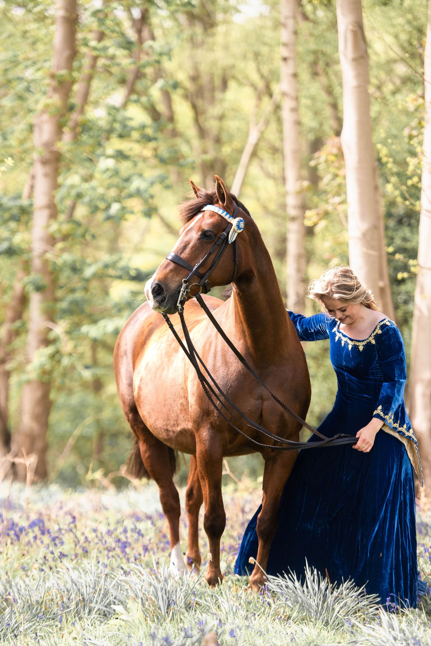 A blonde woman in a blue medieval gown leads a chestnut horse through a bluebell woodland.