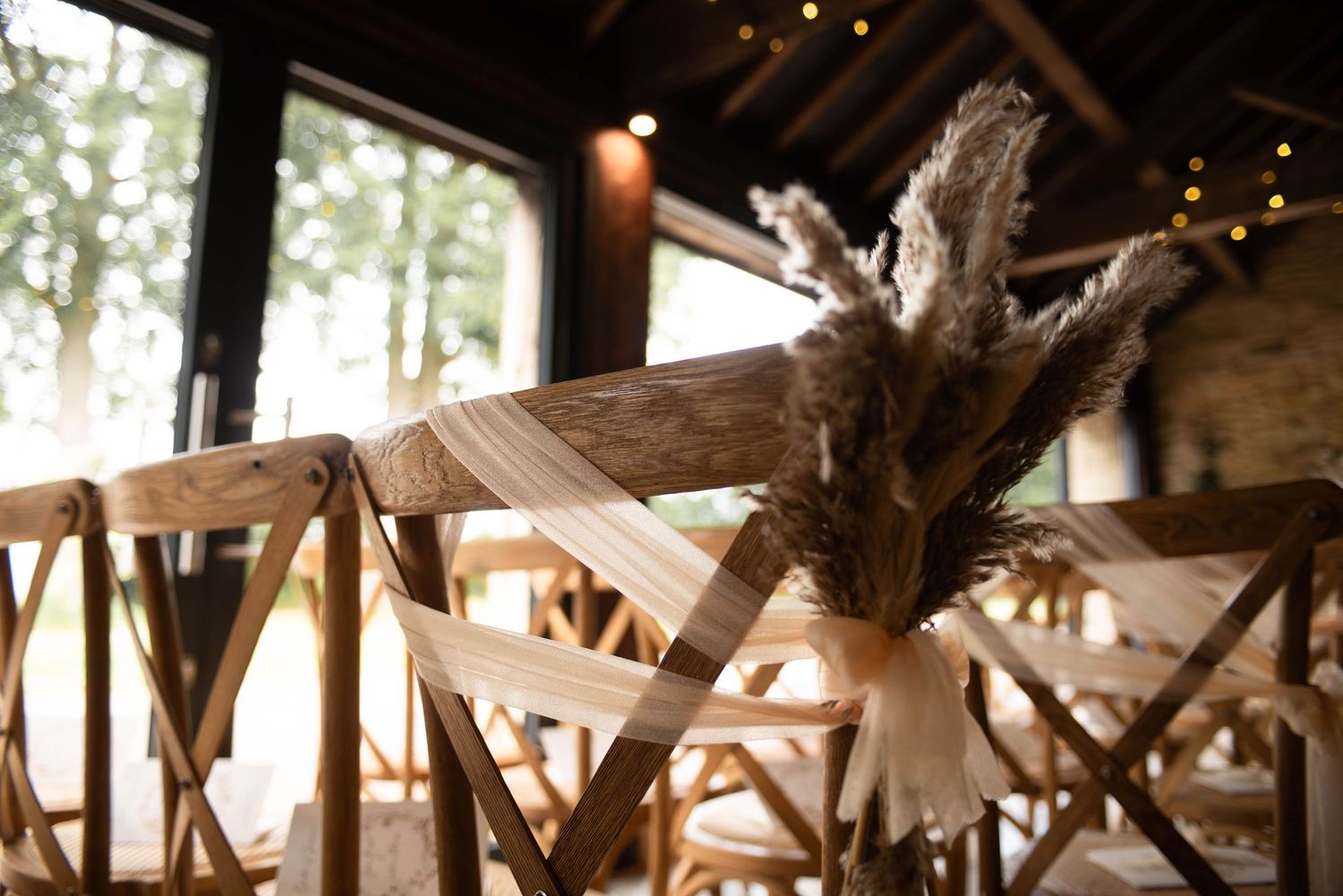 Rustic wooden crossback chairs decorated with pampas grass and tulle in a barn venue.