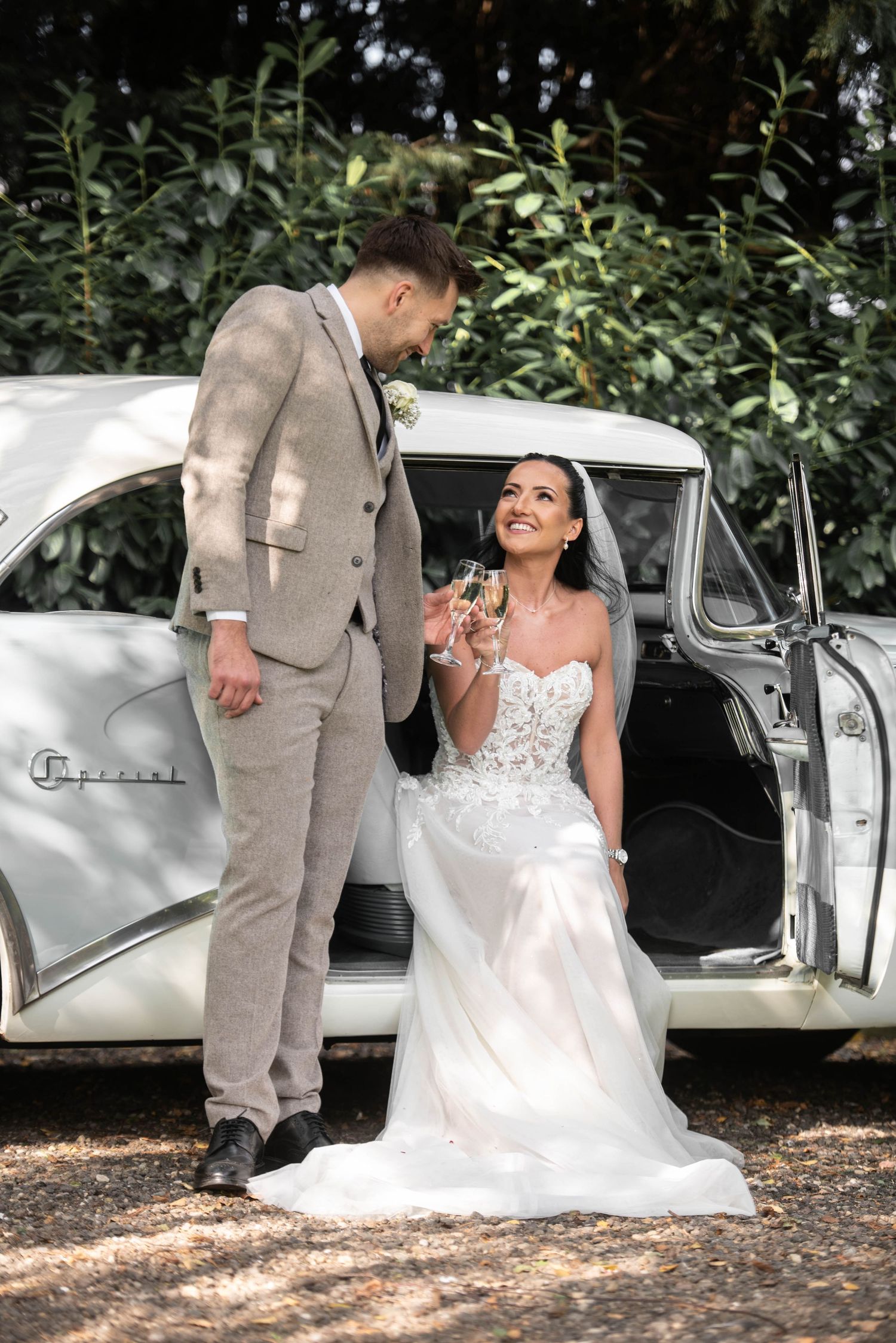 Smiling bride in lace gown holding champagne seated in vintage white car with groom in grey suit.