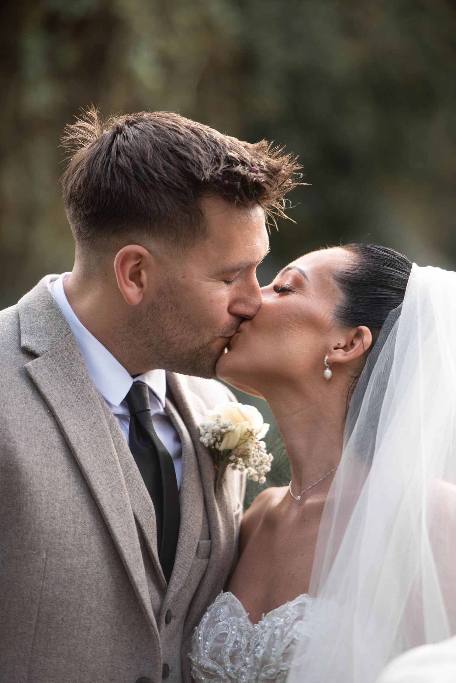 Close-up of bride and groom sharing a kiss, bride wearing a veil and lace gown, groom in grey suit.