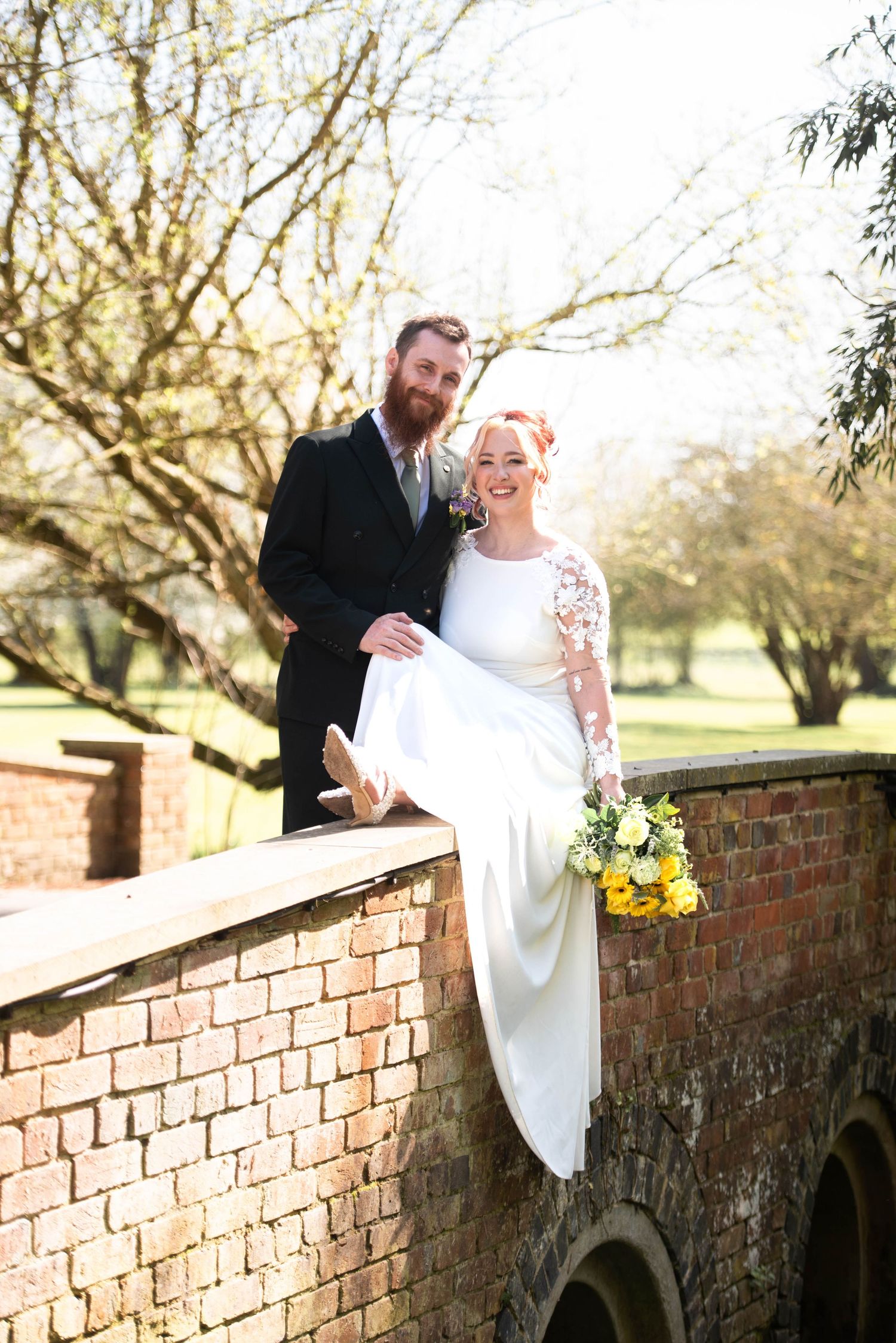 Wedding couple pose on rustic brick bridge, bride in lace gown holding yellow bouquet, groom in dark suit.