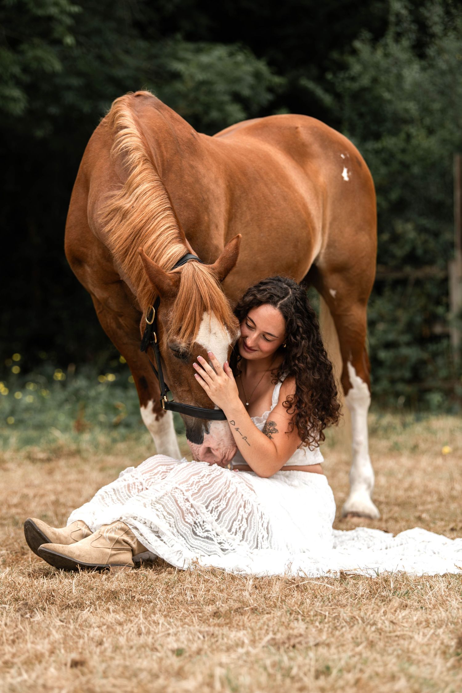 Woman in white dress sitting on ground tenderly petting a chestnut horse in a sunlit field.