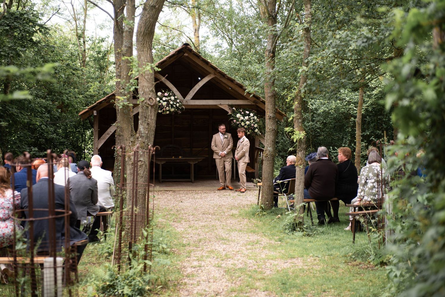 Outdoor woodland wedding ceremony with guests seated along a forest path leading to a rustic wooden arch altar.