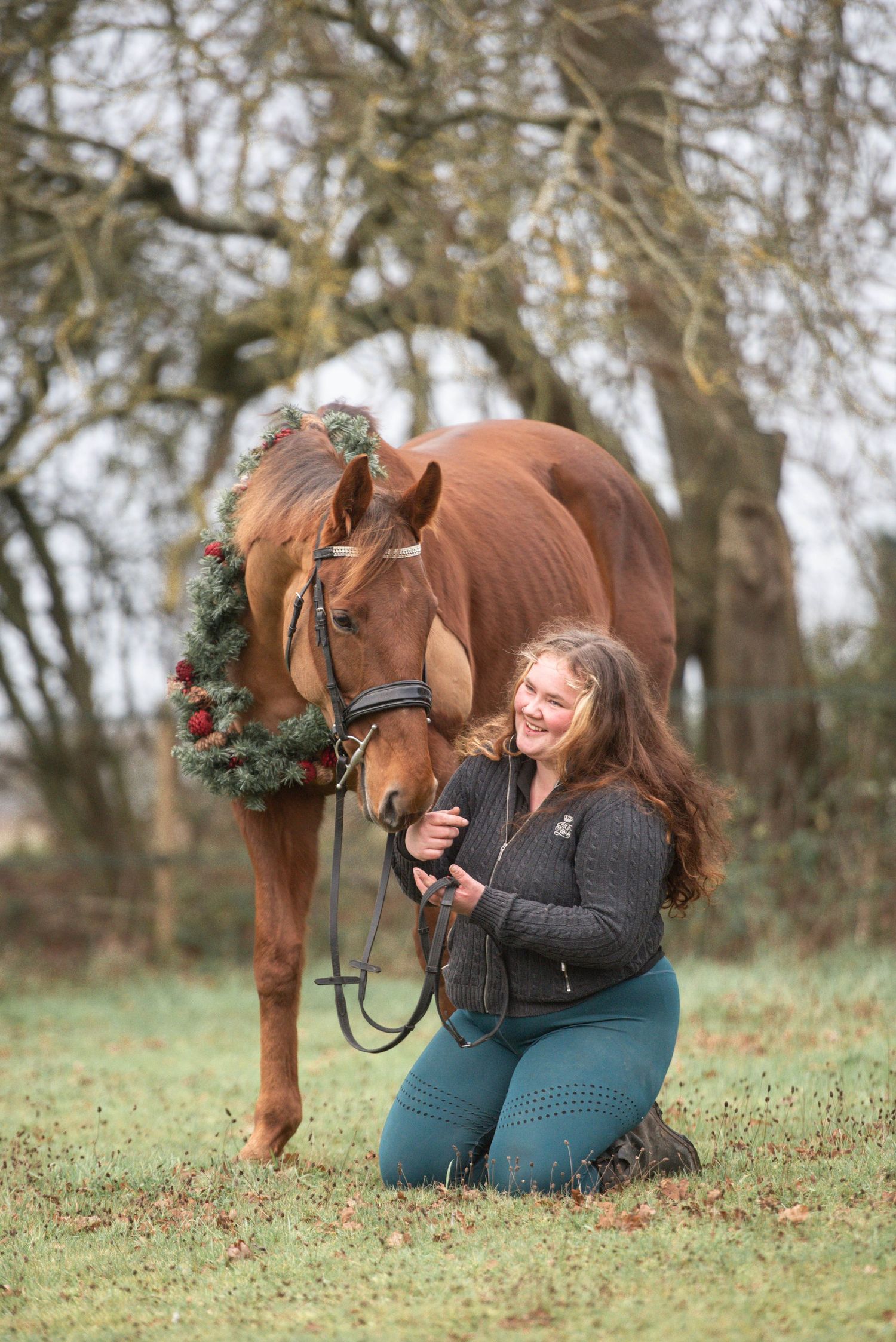 Laughing woman kneeling beside a chestnut horse decorated with a festive Christmas wreath in a field.