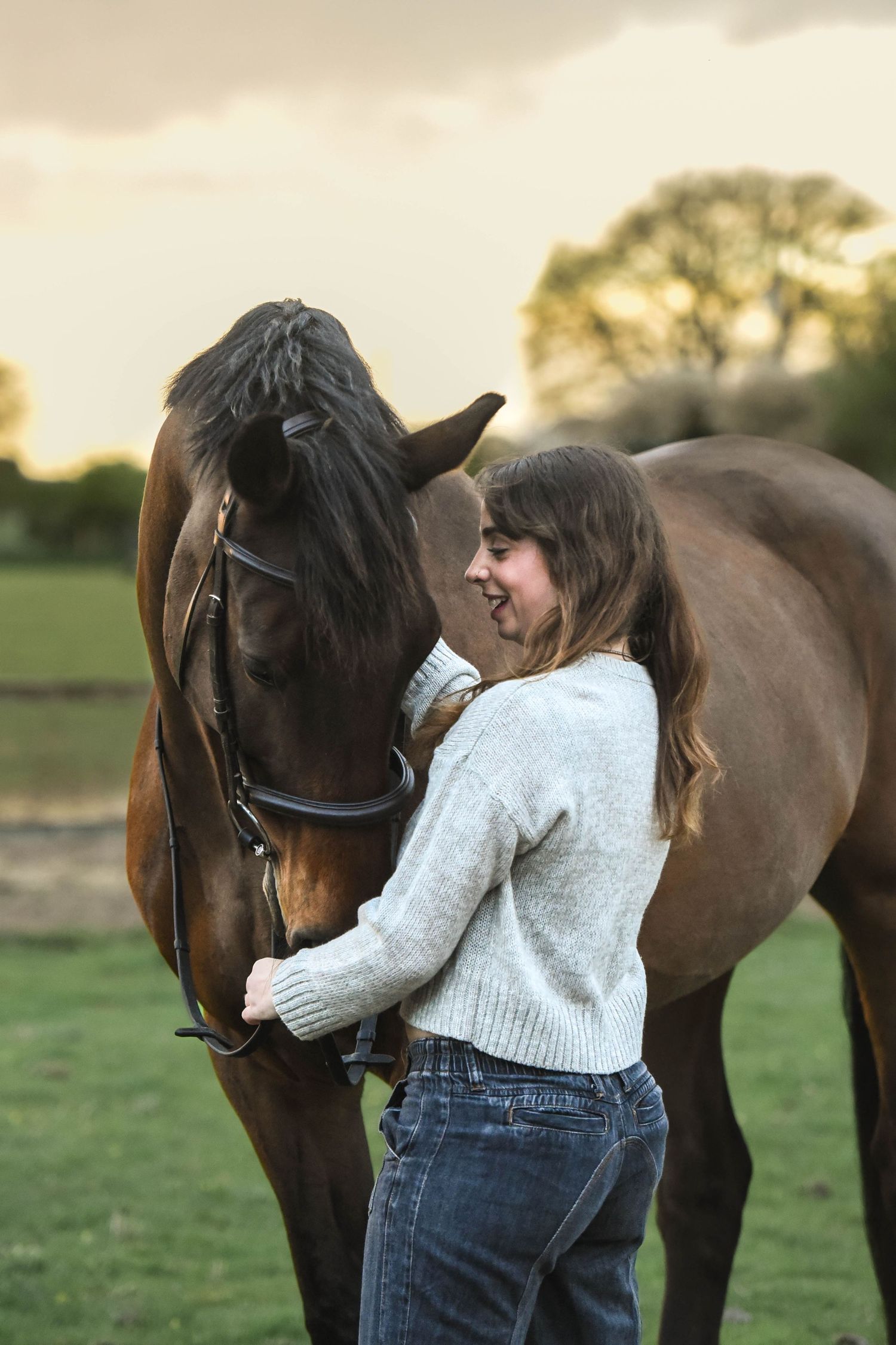 Woman in grey sweater and jeans stands close to a brown bridled horse at dusk outdoors.