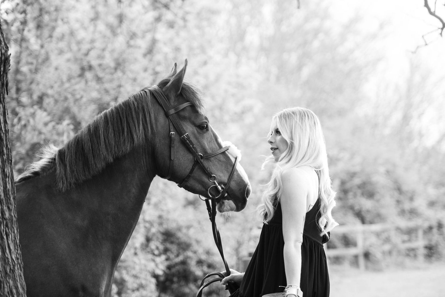 Black and white photo of a blonde woman smiling warmly at a dark horse under leafy trees.