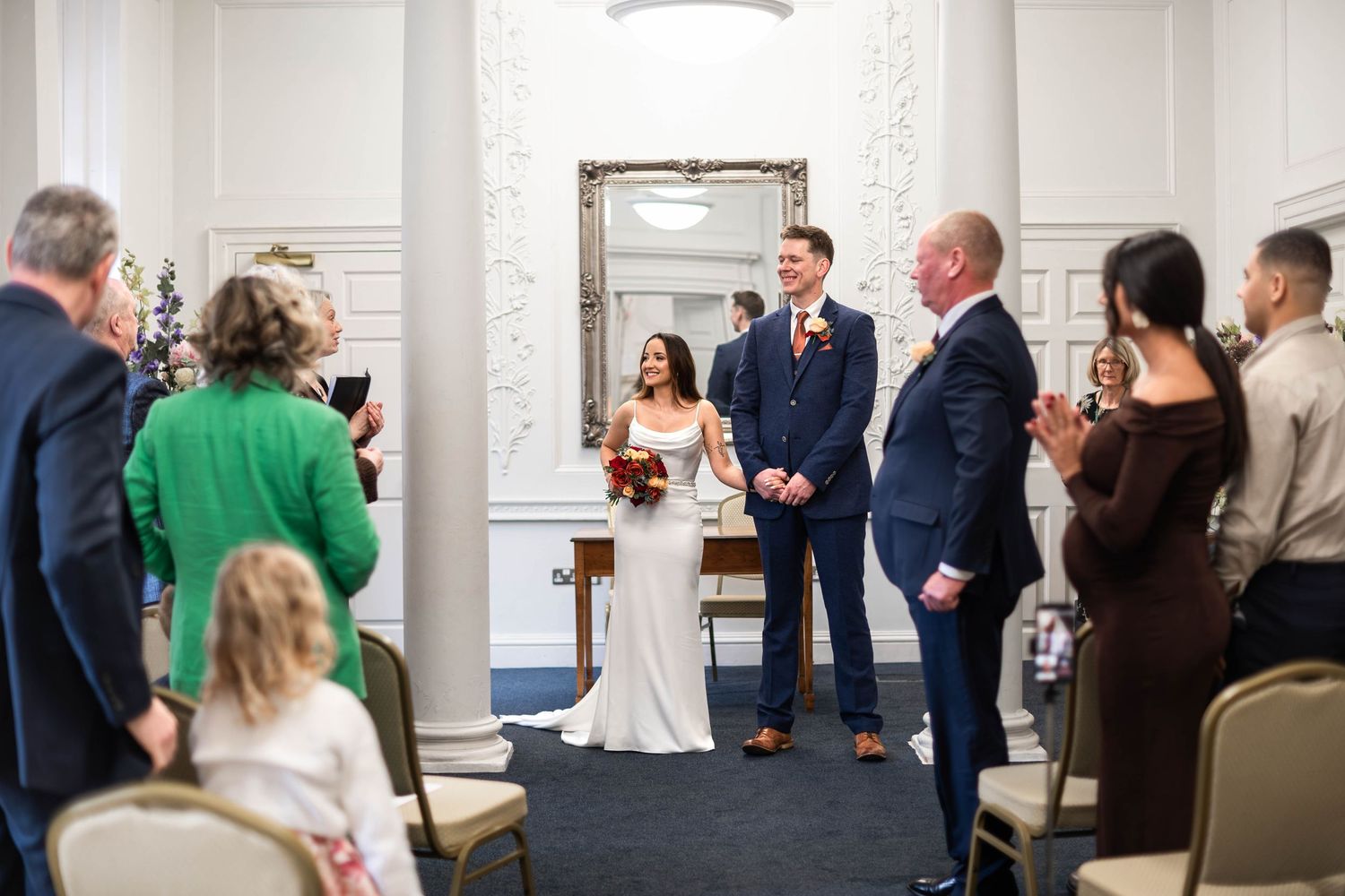 Bride and groom stand together at altar as guests applaud in an elegant white ceremony room.