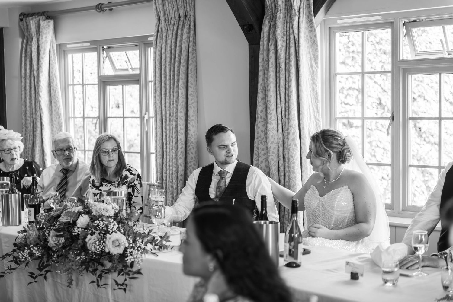 Black and white wedding reception photo of bride and groom seated at table with guests and floral arrangements.