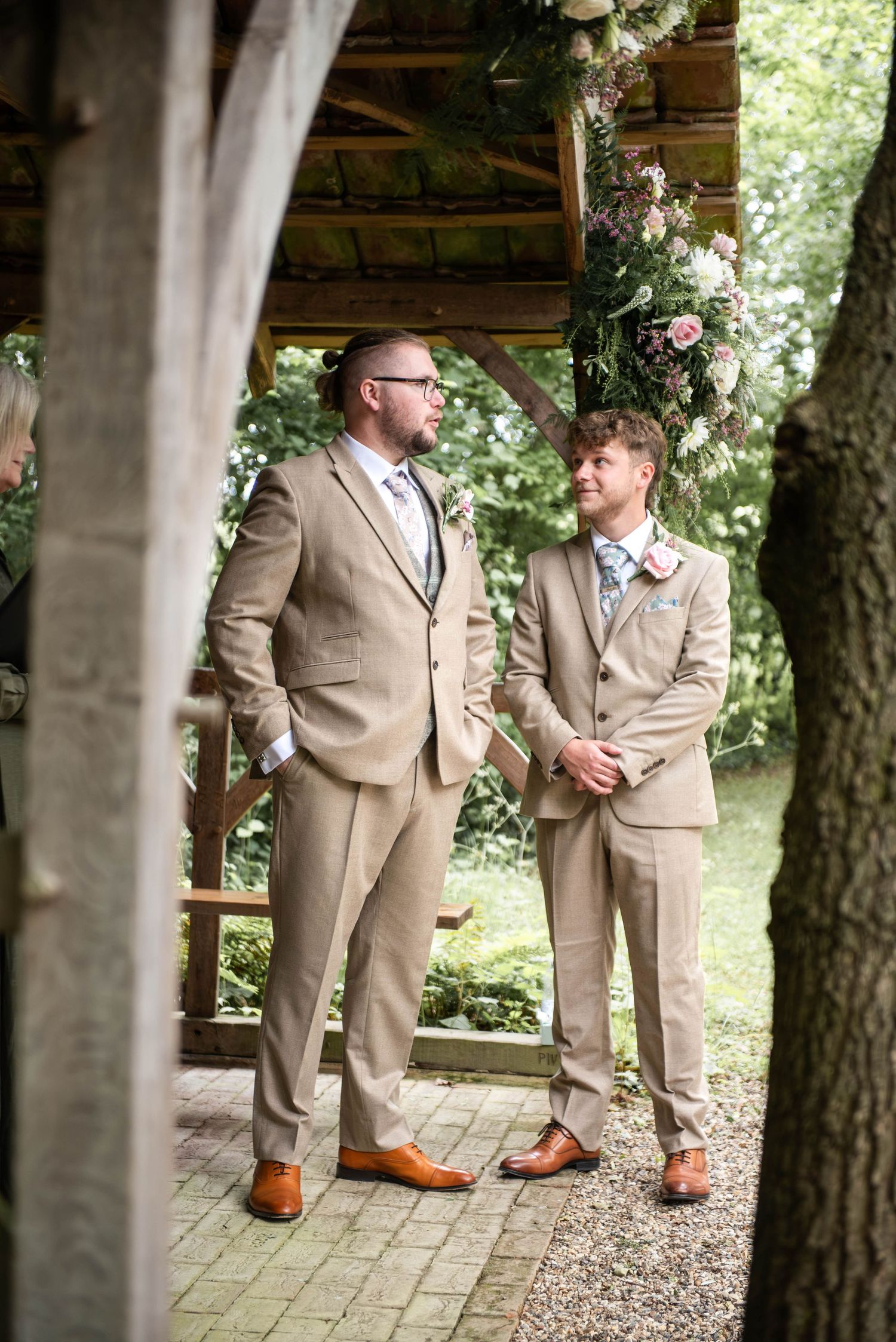 Two grooms in matching tan suits stand together at an outdoor wedding ceremony beneath a floral arch.