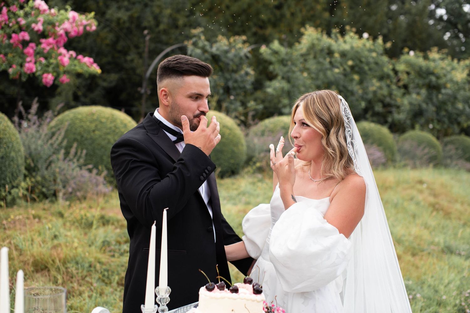 Bride and groom taste wedding cake together outdoors surrounded by blooming roses and garden greenery.