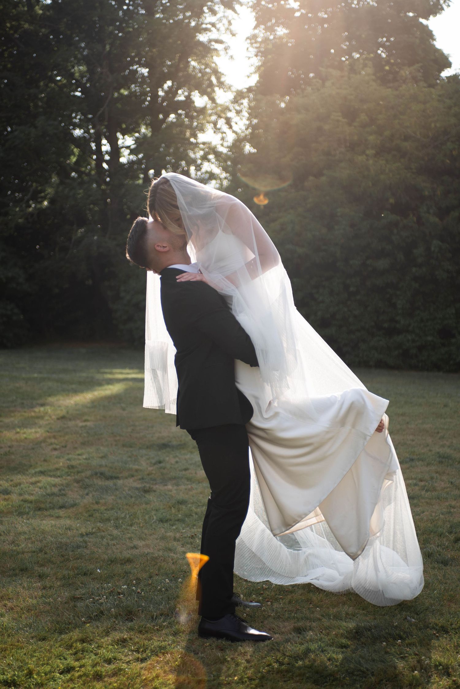 Groom lifts bride in white gown and veil for a romantic kiss in a sunlit garden.
