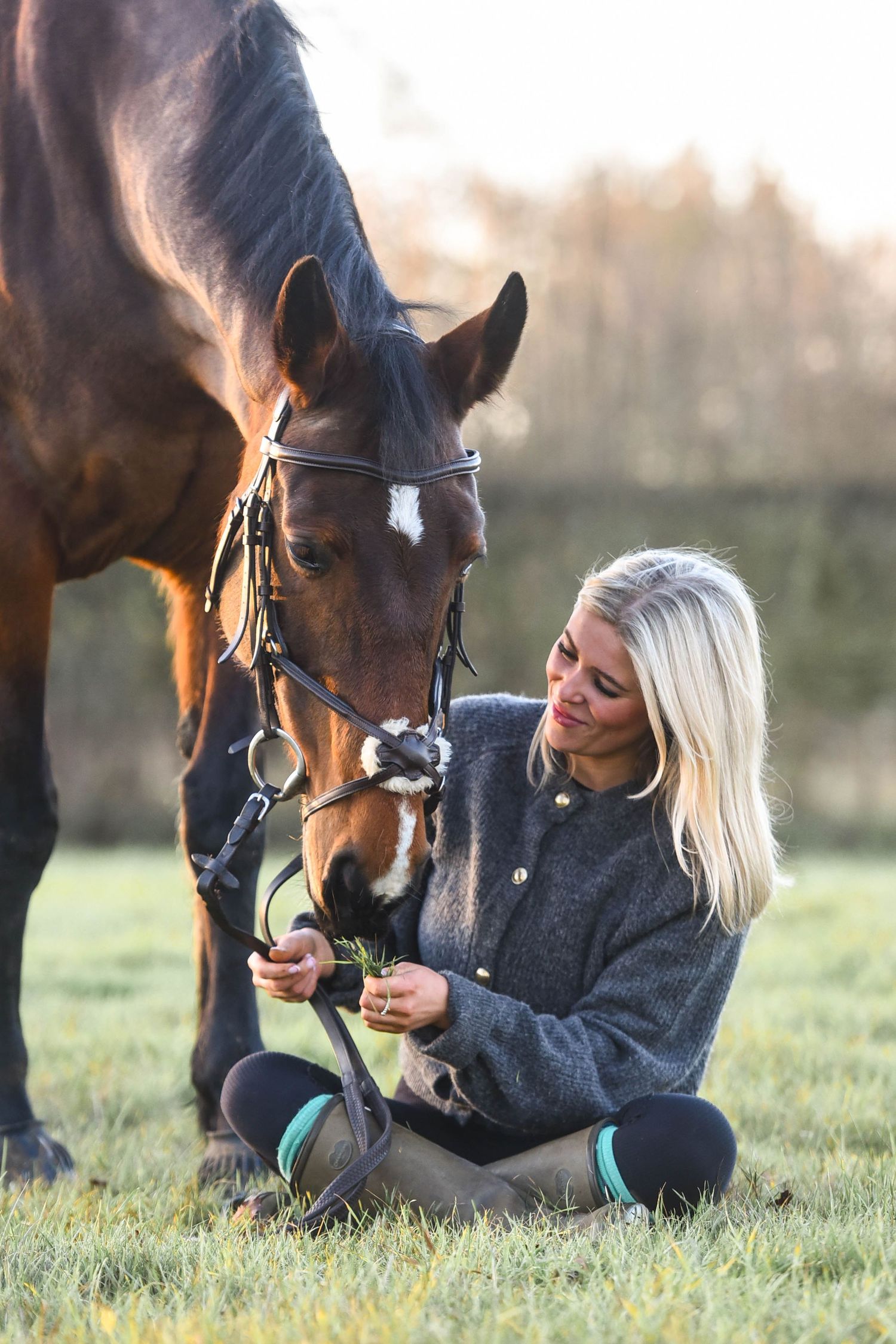Blonde woman sitting on grass feeding a bay horse with a white blaze in golden sunset light.