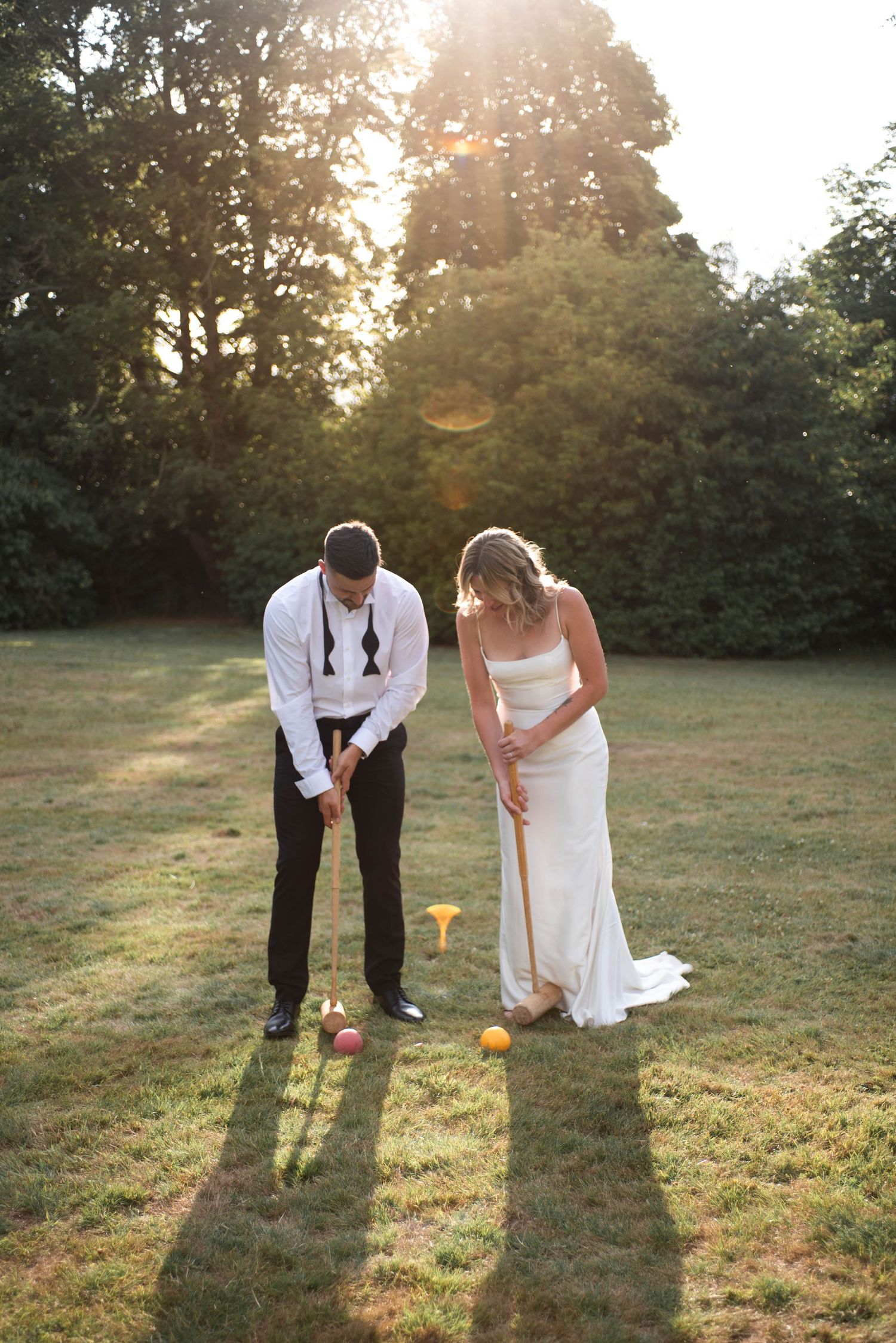 Bride and groom play croquet together on a lawn in warm golden sunset light.