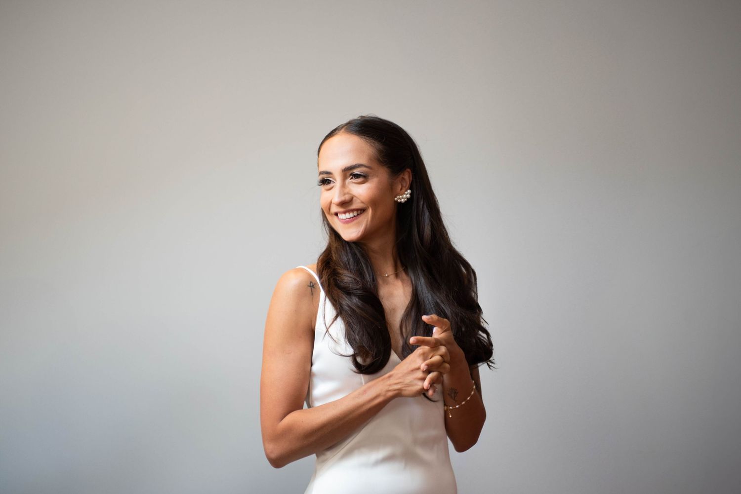 Smiling bride in a white satin slip dress holding her hands together against a neutral background.