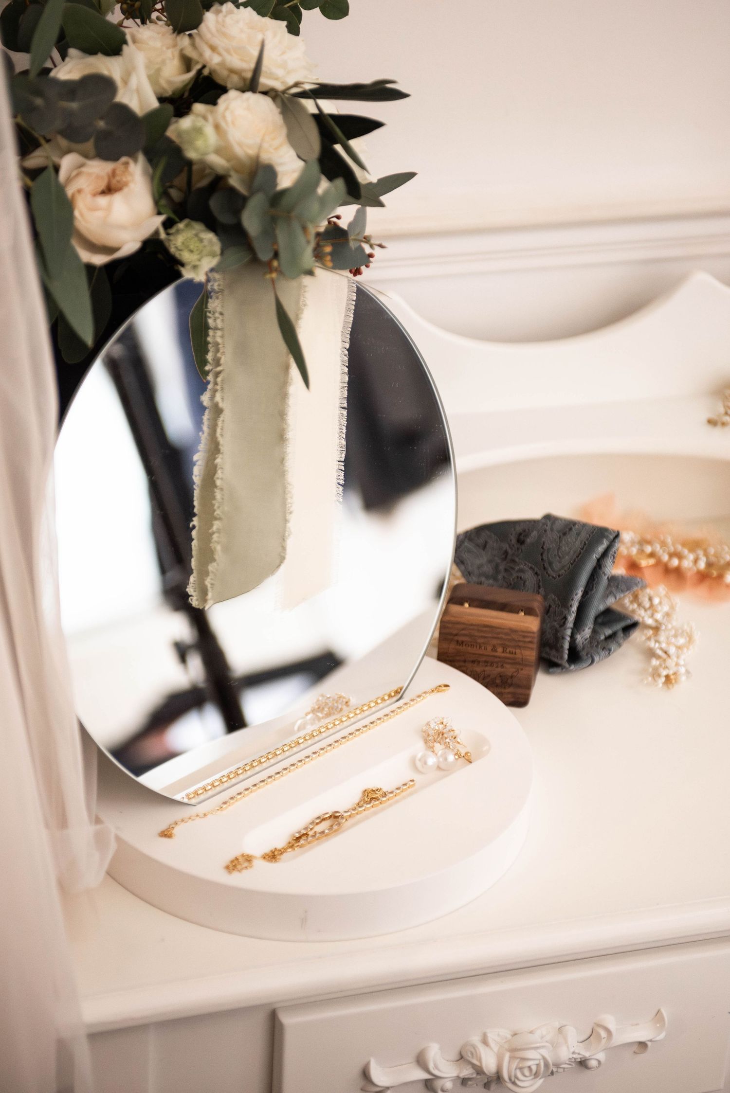 A white vanity dresser displays gold jewelry and white roses reflected in a round mirror.