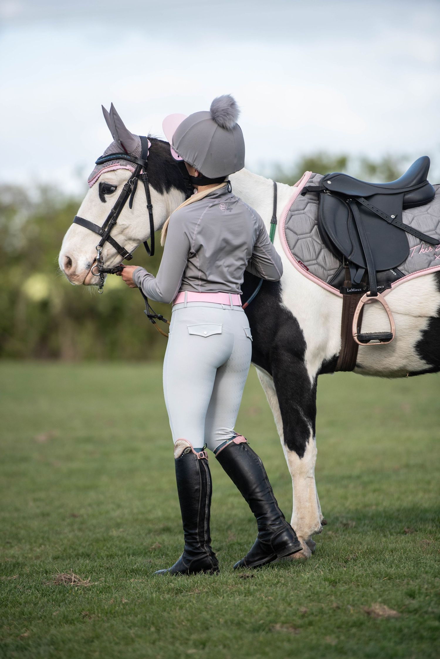 Young equestrian in pink and grey riding gear standing with black and white horse.