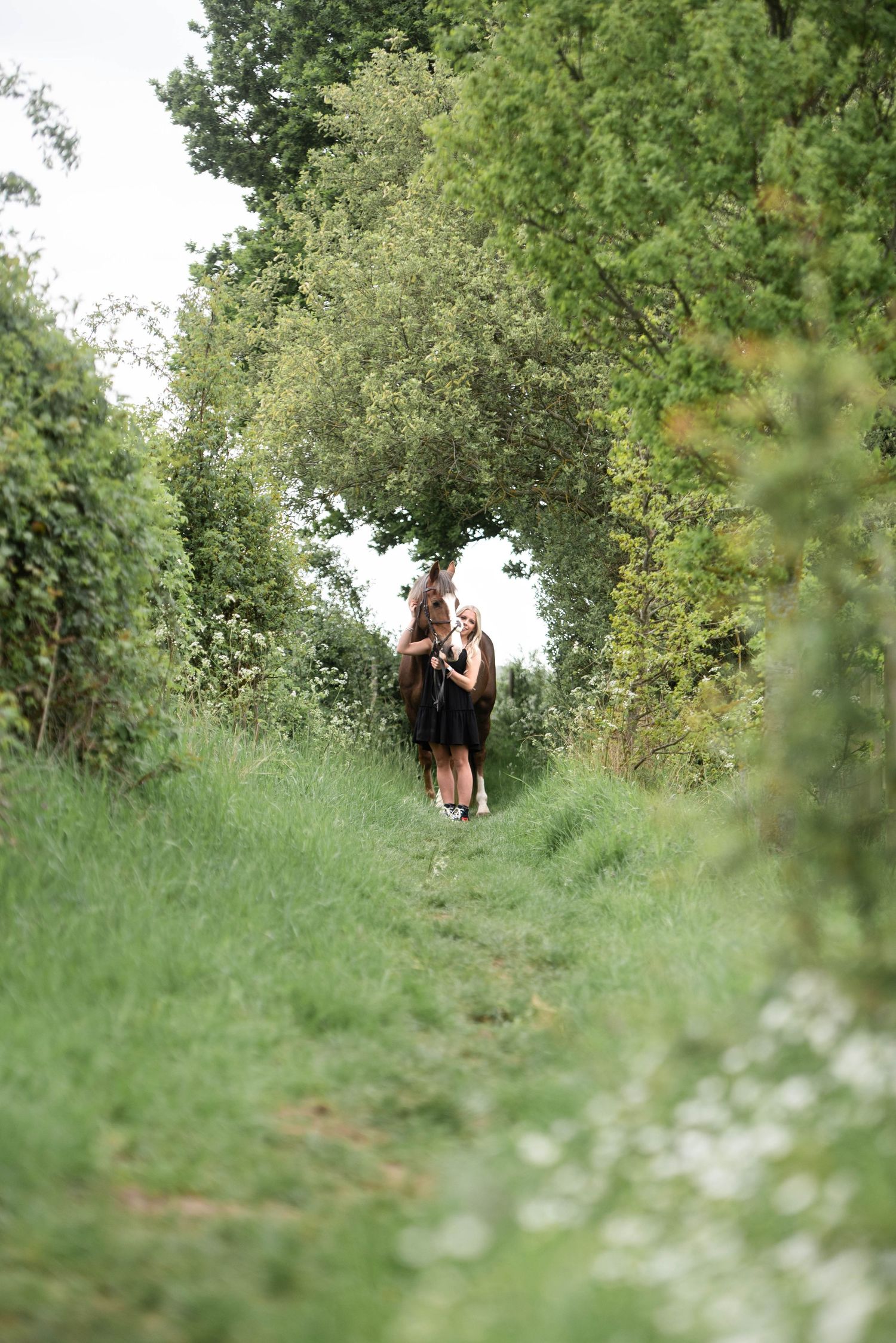 Woman in black dress leading a chestnut horse along a lush green tree-lined path.