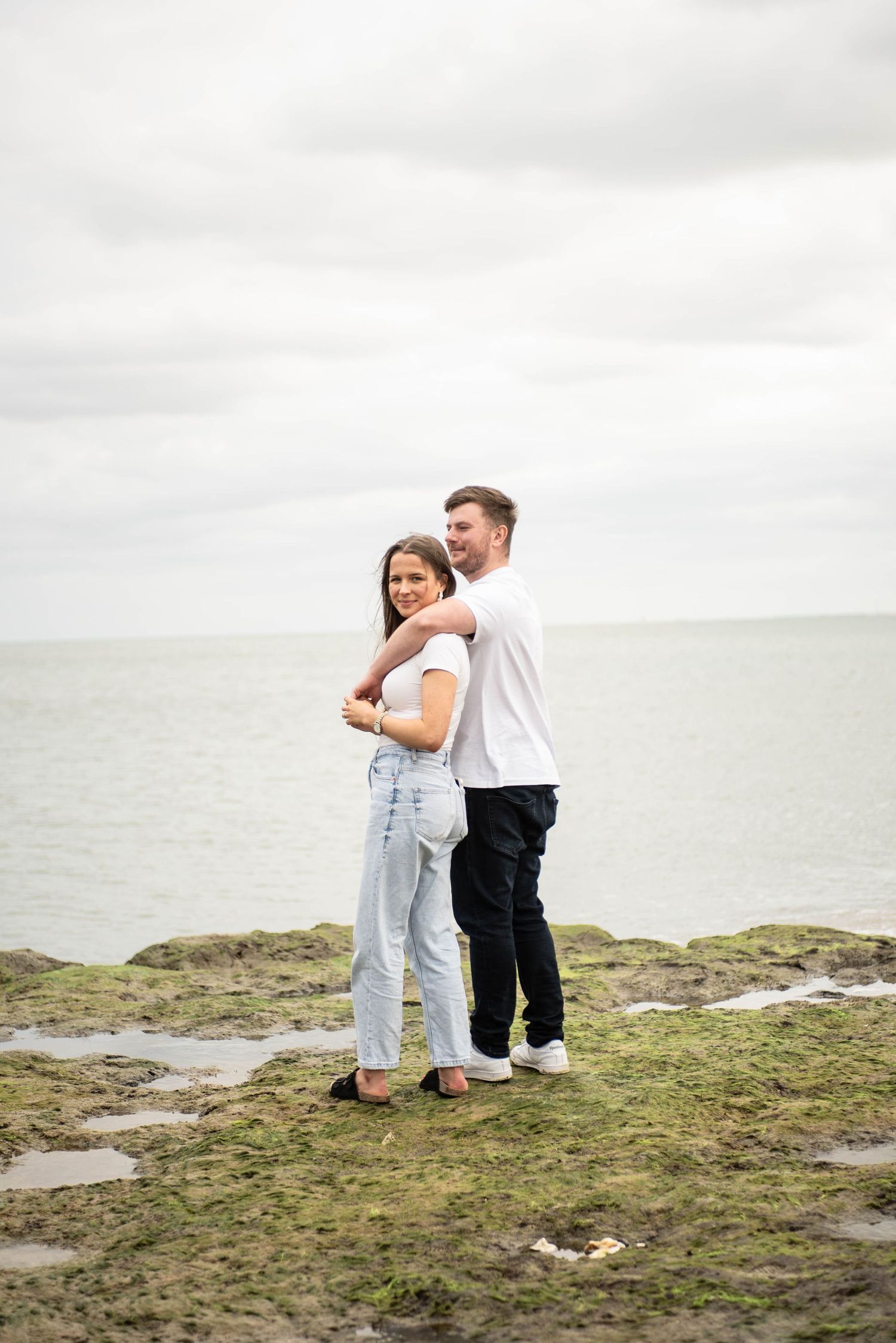 A young couple embracing on a rocky mossy coastline under an overcast sky.