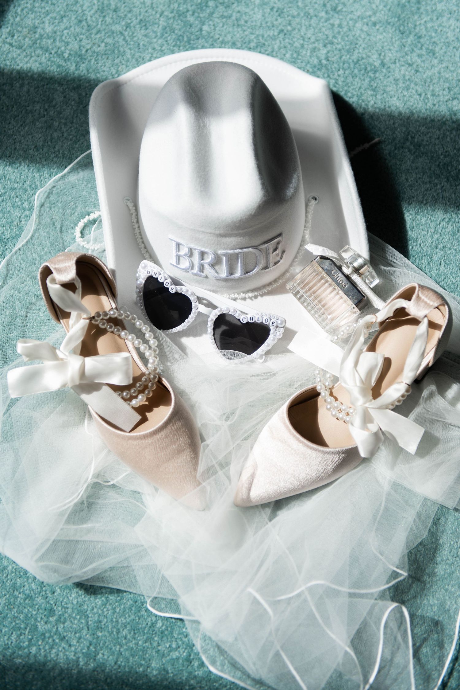 Bridal flat shoes with bow details, heart-shaped sunglasses, and a white cowgirl hat labeled BRIDE on tulle veil.